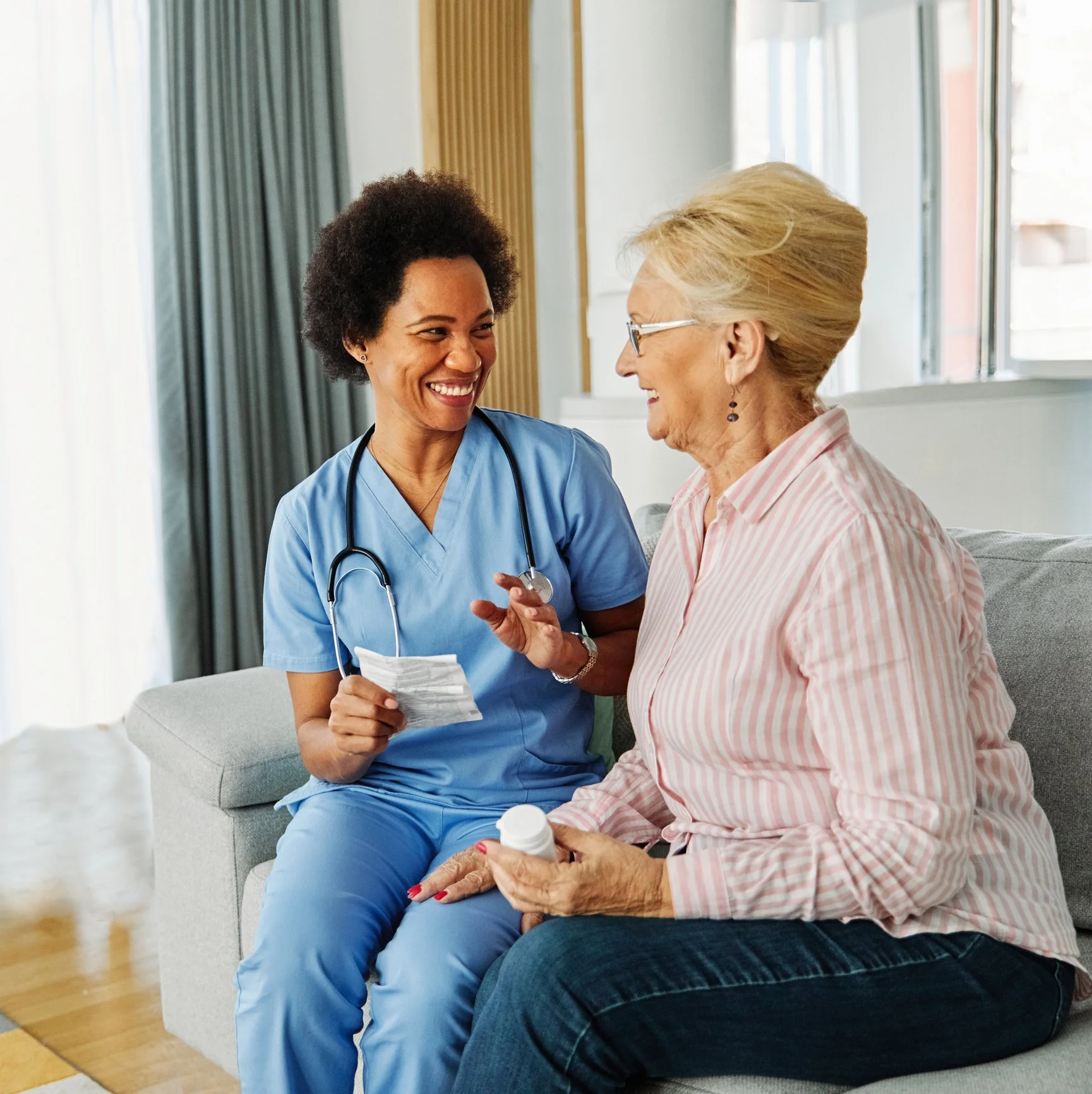 A healthcare worker and an elderly woman sharing a joyful moment on a sofa indoors, with the woman holding a bottle.