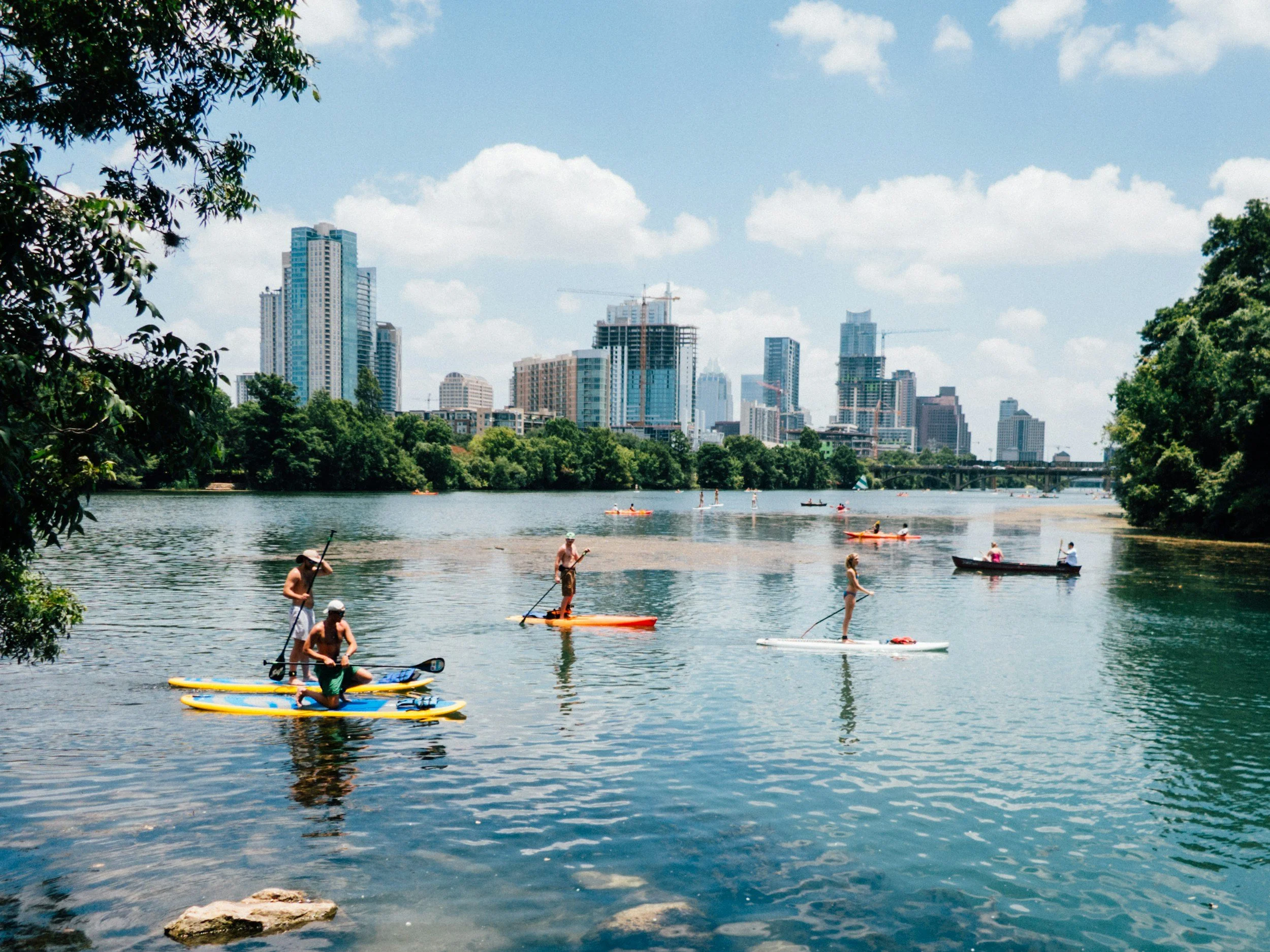 People paddleboarding and kayaking on a calm river with a city skyline in the background on a sunny day.