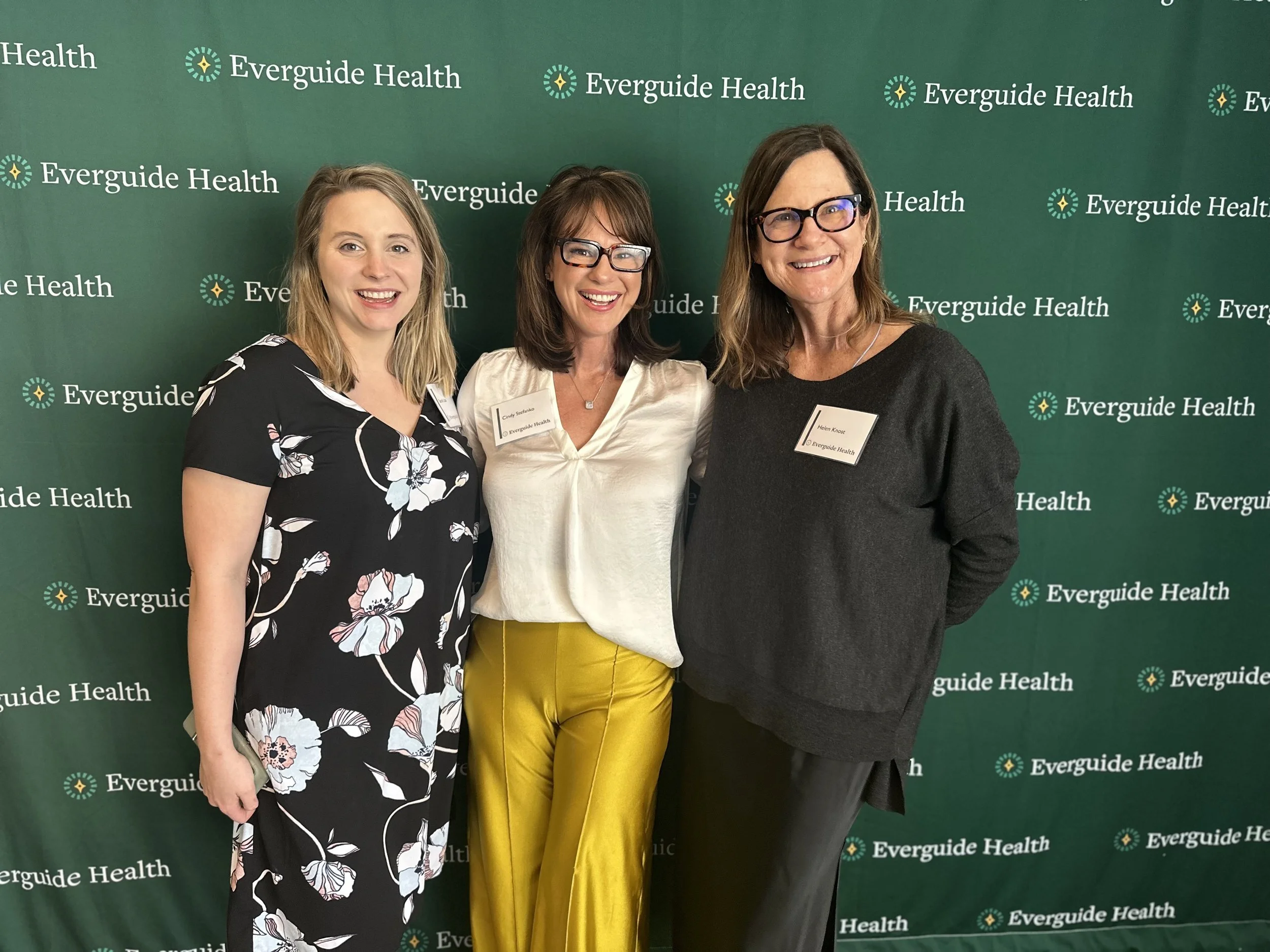 Three women standing together in front of a green backdrop with 'Everguide Health' written repeatedly. They are smiling and dressed in business casual attire; the woman on the left wears a black floral dress, the woman in the middle wears a cream-colored blouse and yellow pants, and the woman on the right wears a dark gray sweater and black pants.