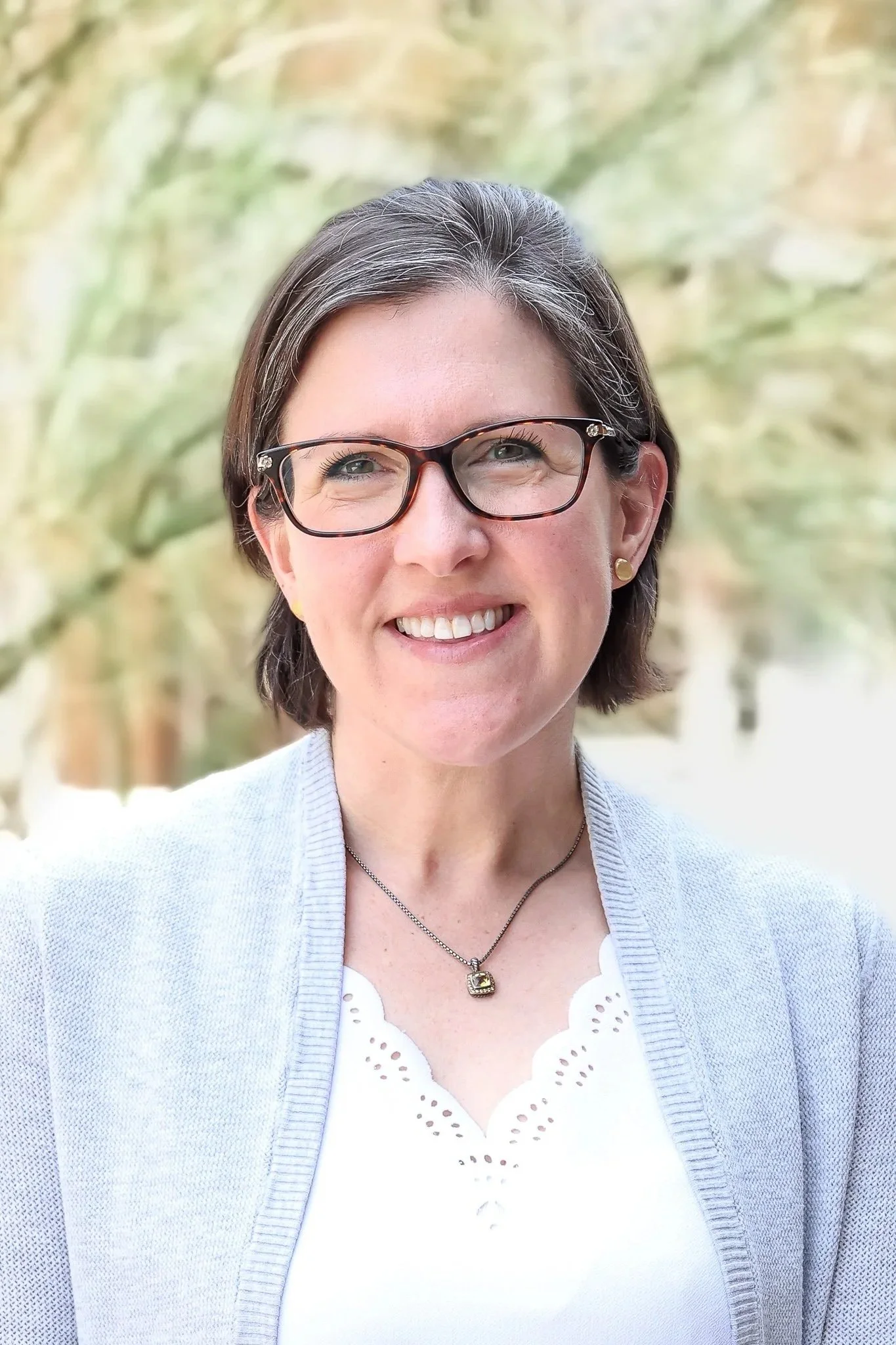 Portrait of a woman with short dark hair, glasses, wearing a white top, light gray cardigan, a necklace, and earrings, smiling outdoors with blurred trees in the background.