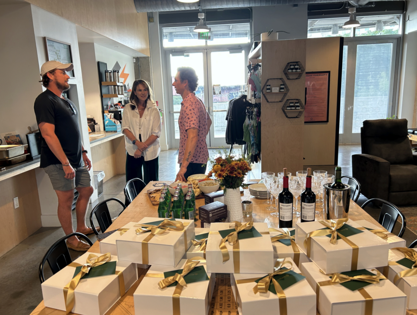 Three people standing and talking in a cafe or event space, with a table of gift boxes, drinks, and food in the foreground.