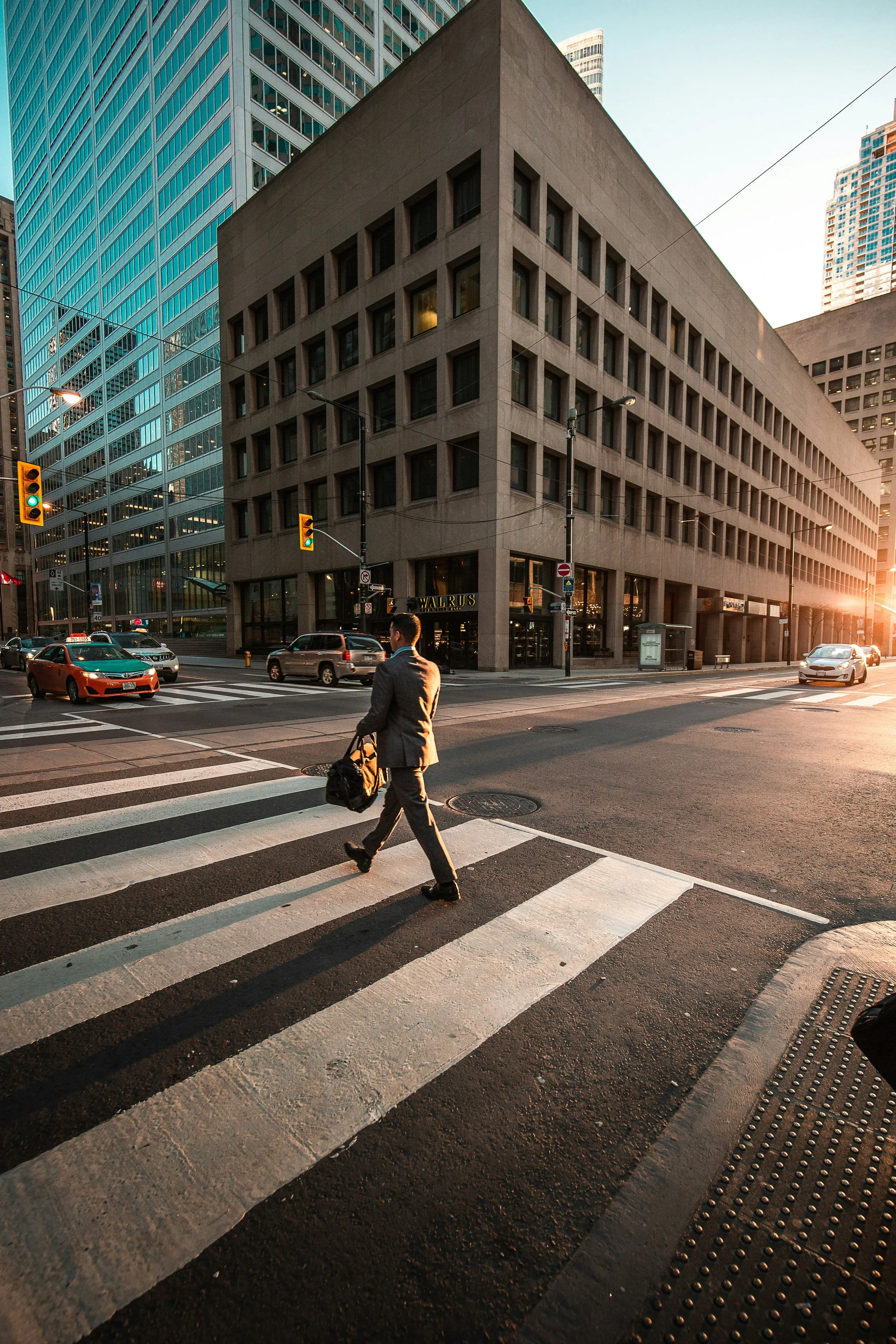 A man in a business suit crossing a city street at sunset, carrying a bag, with tall modern buildings and traffic around him.