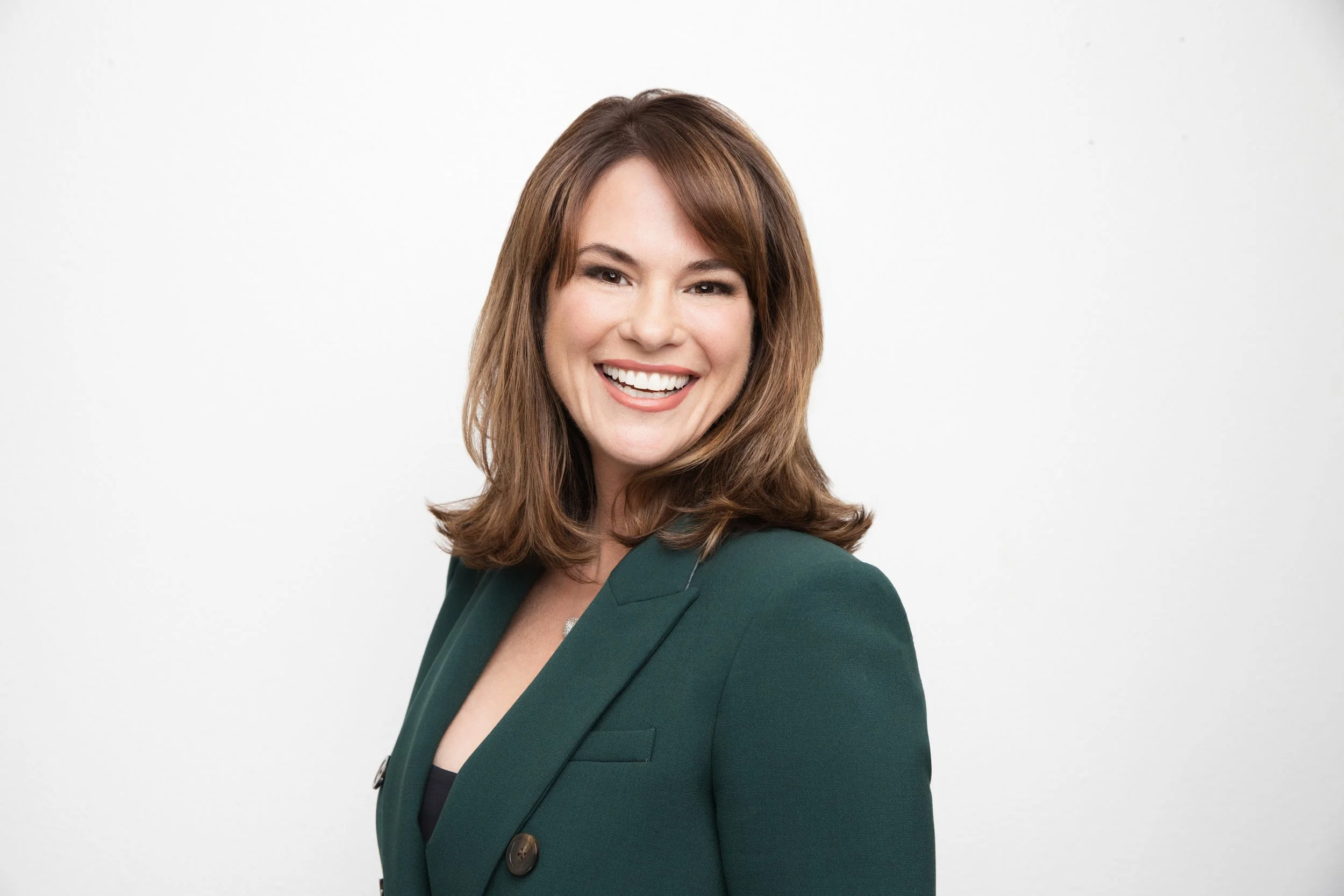 A woman with shoulder-length brown hair, wearing a dark green blazer, smiling against a plain white background.