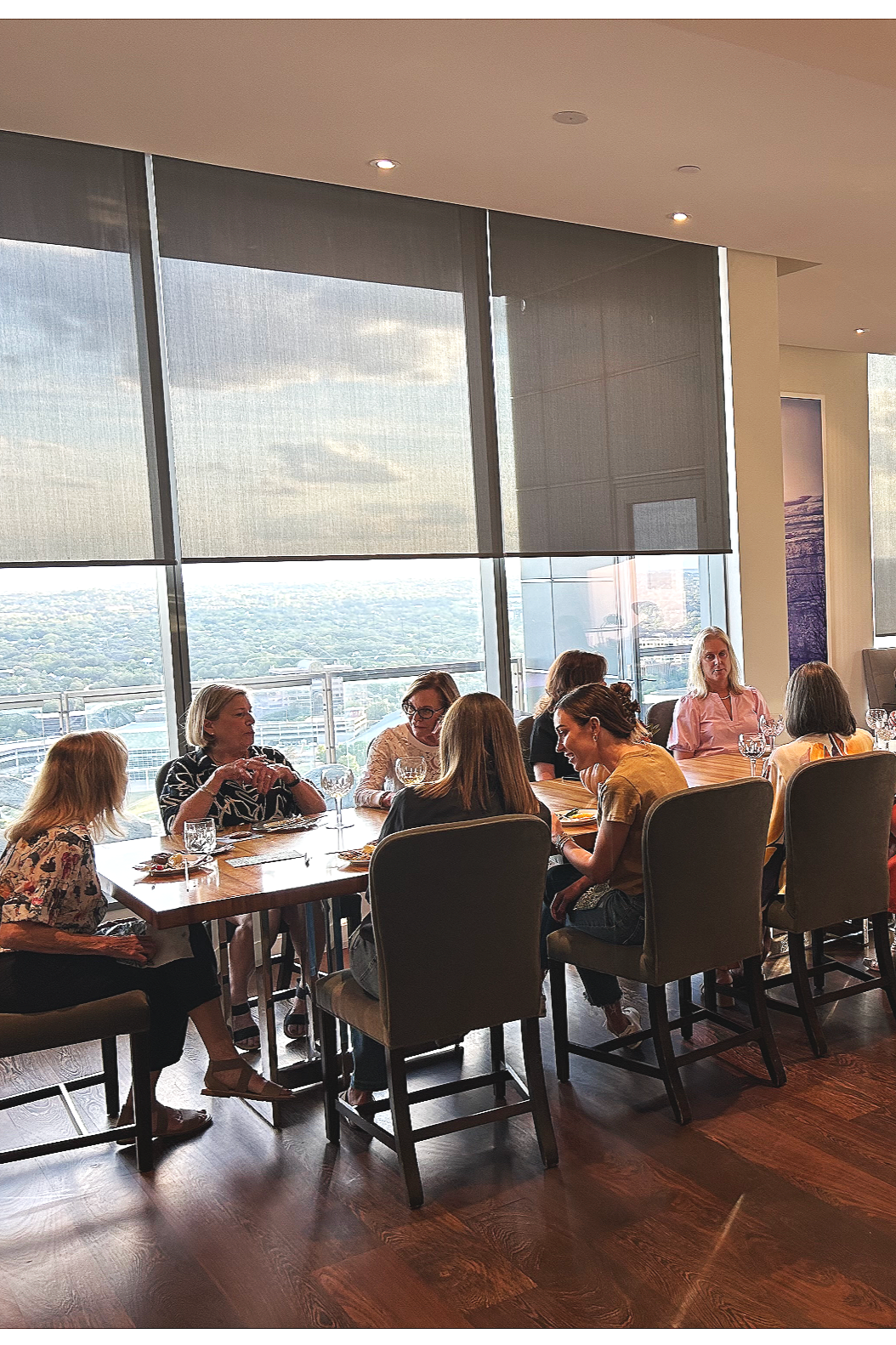 A group of women sitting around a dining table in a high-rise restaurant with large windows showing a cityscape and cloudy sky, engaged in conversation.