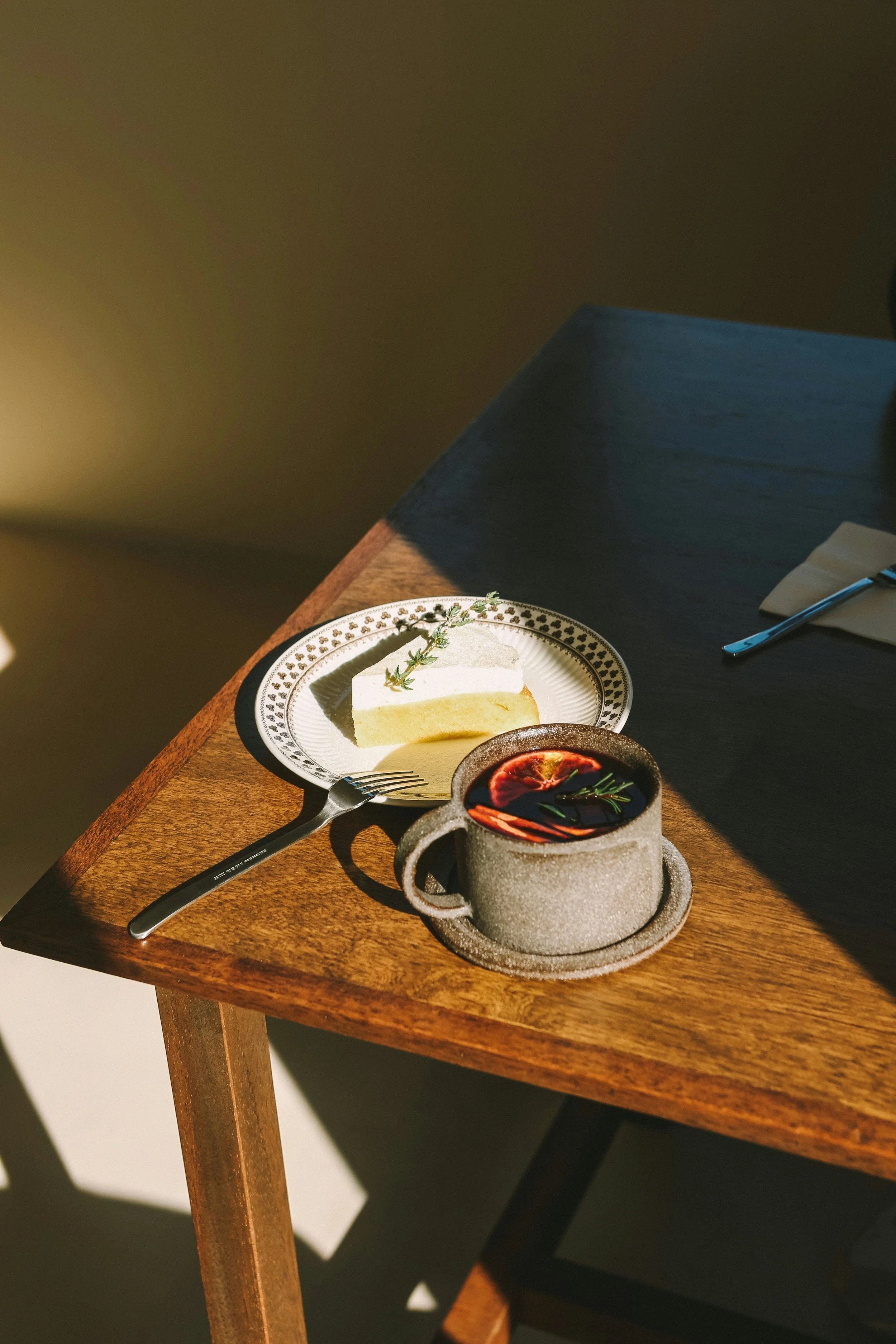 A slice of cake and a cup of dark beverage with slices of fruit and herbs on a wooden table.