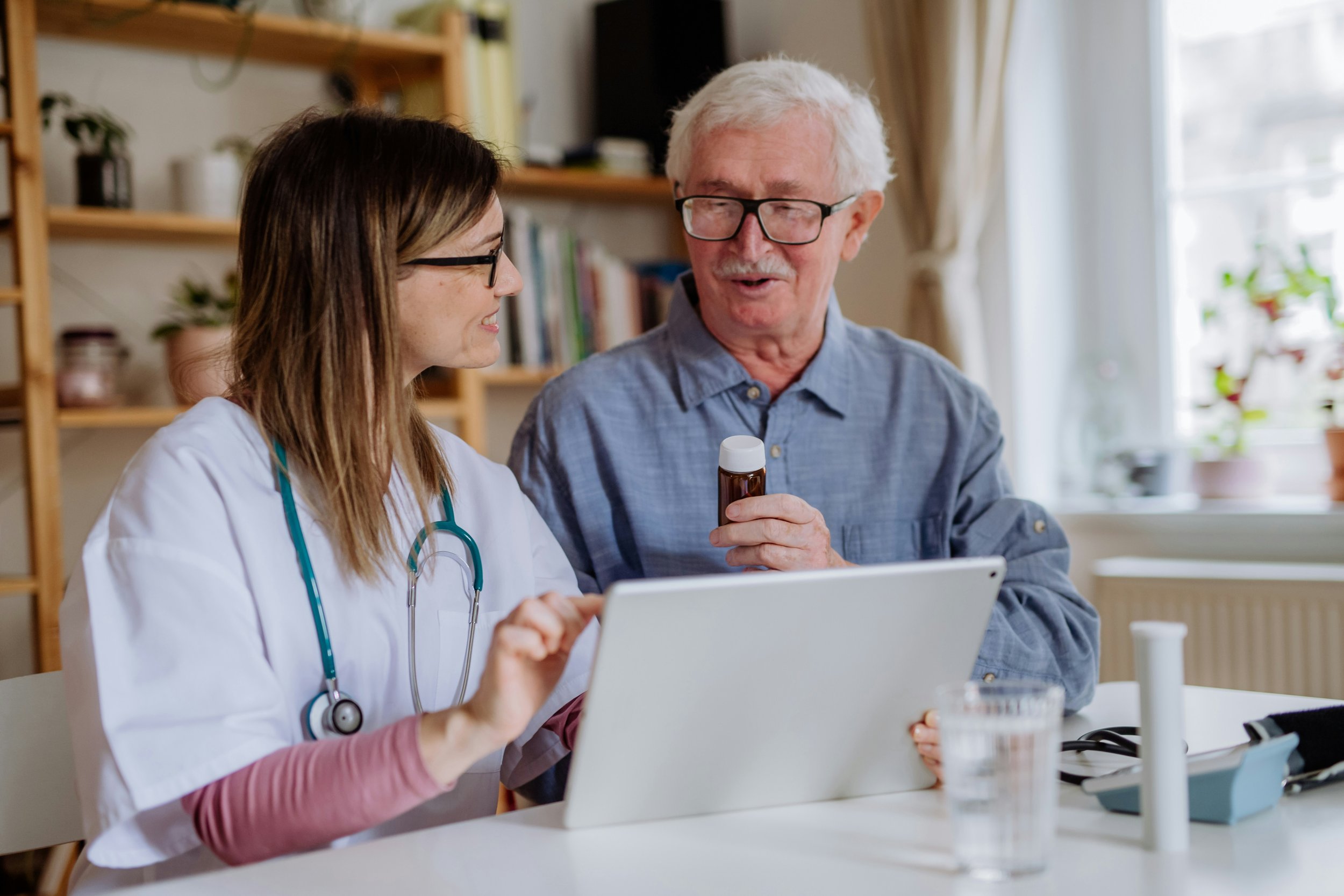 A young female healthcare worker with a stethoscope around her neck showing a tablet to an elderly man holding a medication bottle in a well-lit room with bookshelves and plants.