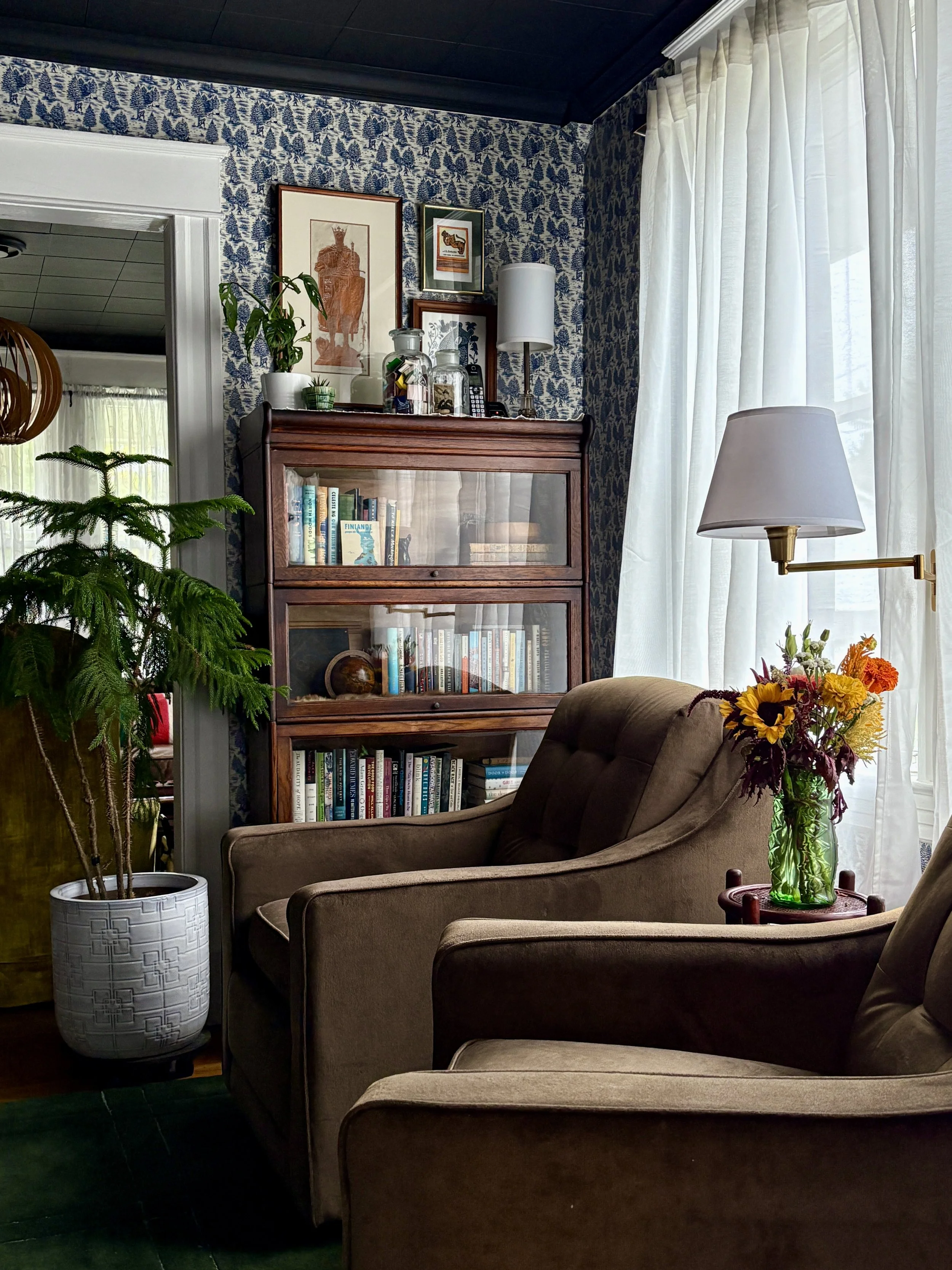 A cozy living room corner with a brown armchair, a side table with a vase of colorful flowers, a floor lamp next to a window with white curtains, a wooden bookshelf filled with books, decorative glass jars and plants, and a background of blue patterned wallpaper and framed artwork.