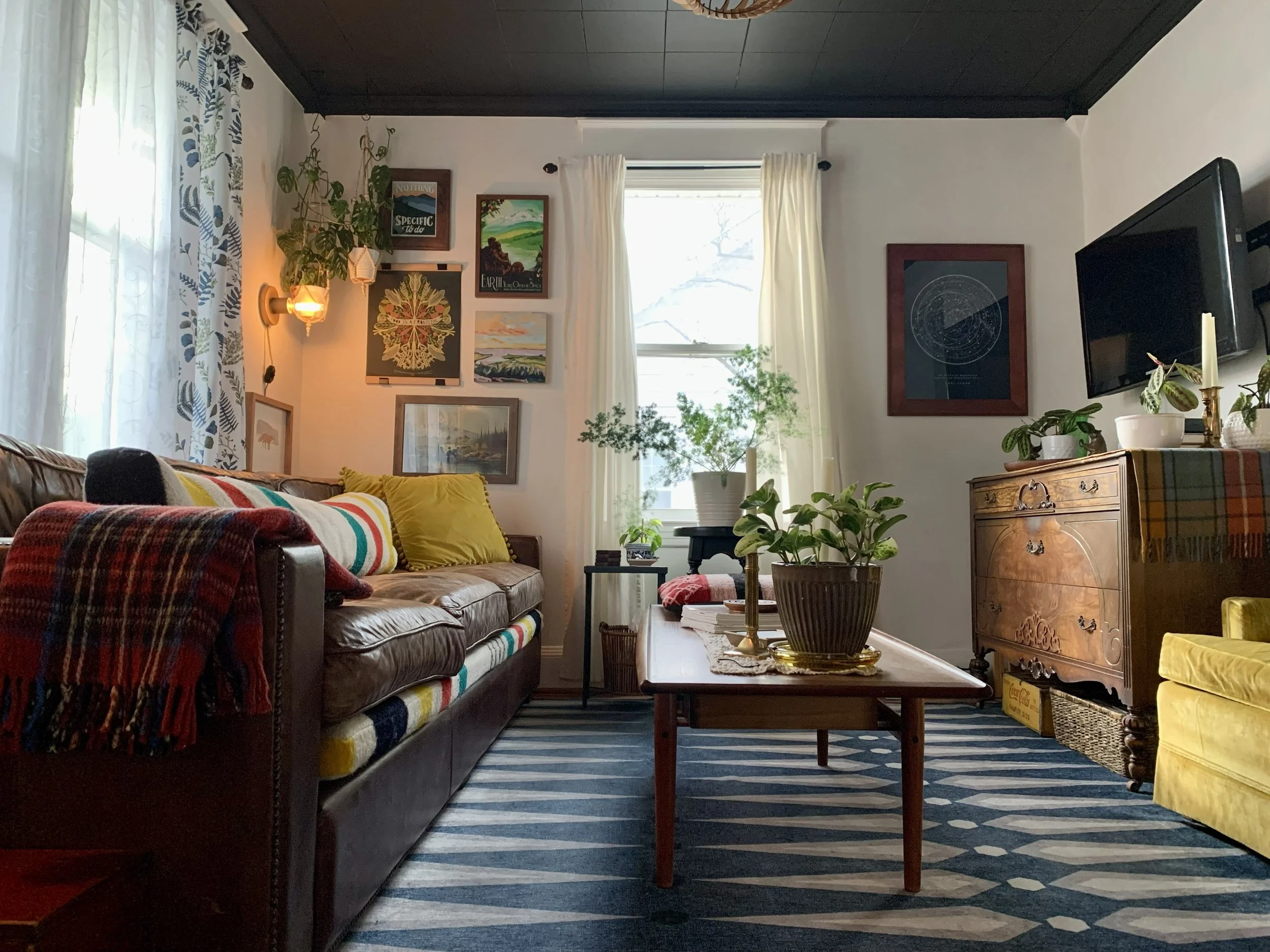 Living room with a leather sofa with colorful cushions and a plaid blanket, a wooden coffee table with potted plants, a yellow armchair, a wooden sideboard with potted plants and a framed artwork, a wall with various framed pictures, and a window with light curtains.