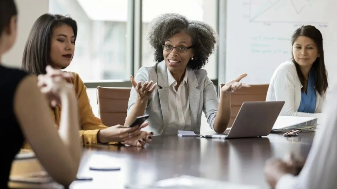 A diverse group of women in a business meeting, with one woman speaking animatedly while others listen and engage.