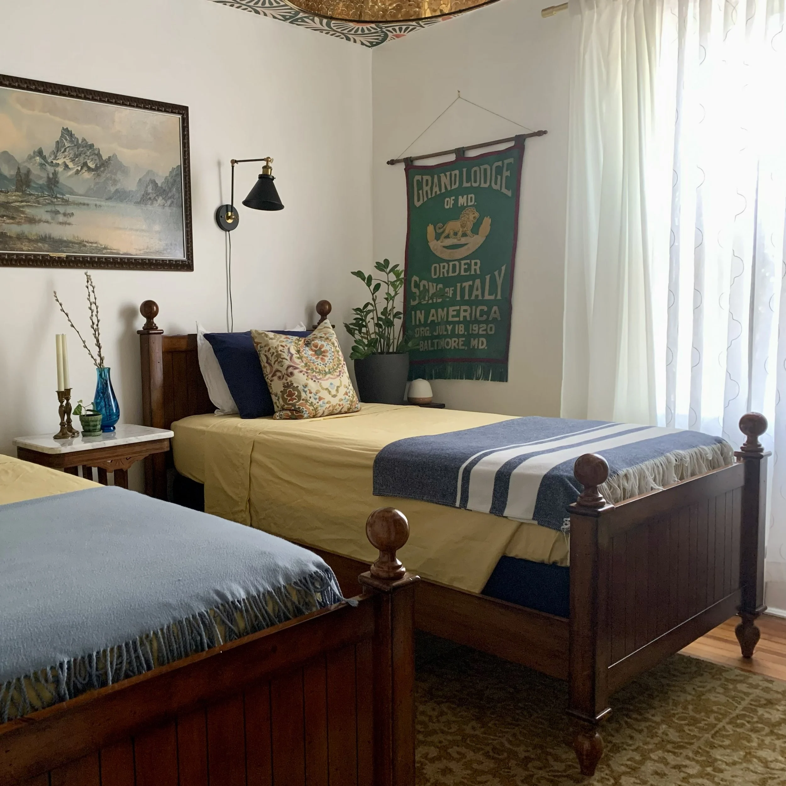 A bedroom with two wooden beds, one with a yellow bedspread and the other with a blue and white striped blanket, a nightstand with decor, a hanging green banner on the wall, a window with sheer curtains, and a framed landscape painting, in a vintage style.