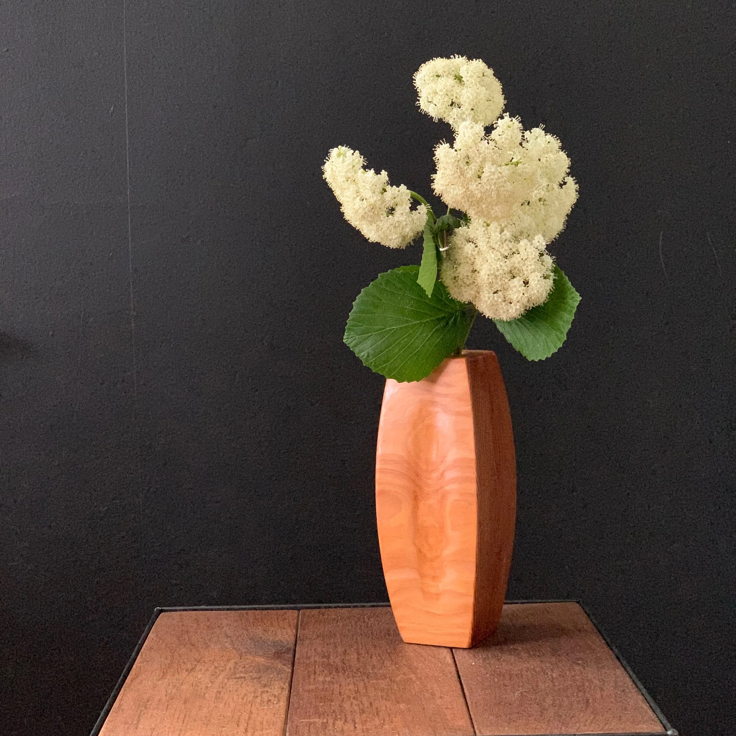 A wooden vase with white hydrangea flowers and green leaves against a black background.