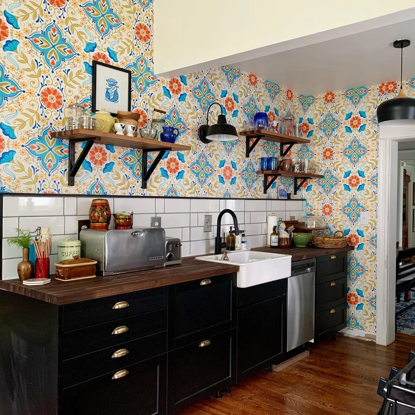 A kitchen with a colorful floral patterned wallpaper, black cabinets, wooden countertops, white tile backsplash, open wooden shelves with dishware and decorative items, a farmhouse sink, and a stainless steel dishwasher.