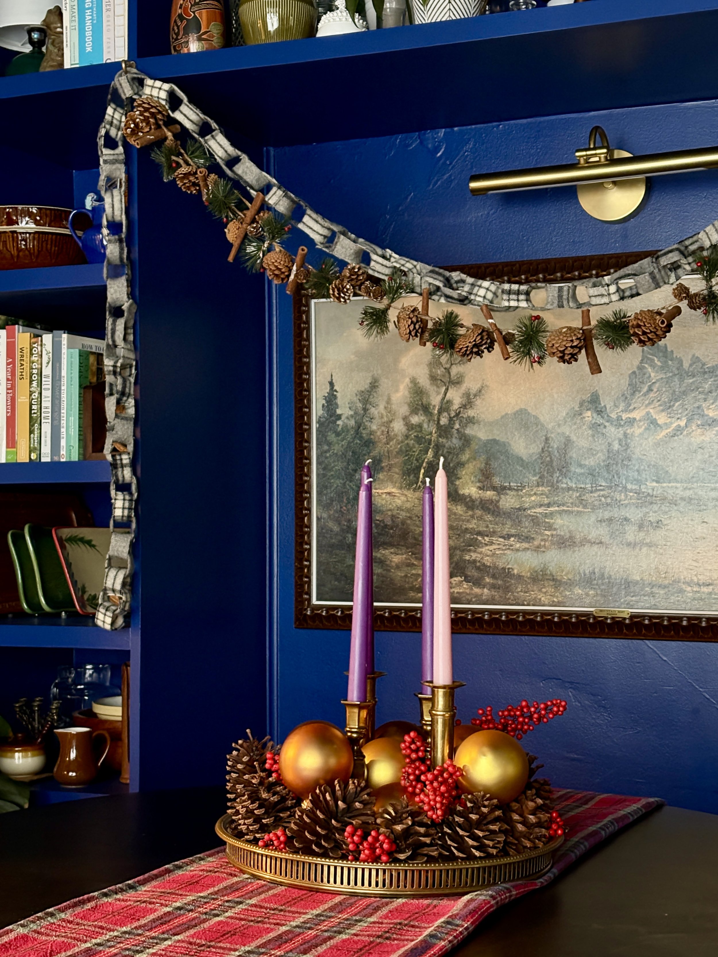 A festive holiday centerpiece with purple and pink candles, gold ornaments, pinecones, and red berries on a red plaid table runner in a room with blue walls, a framed landscape painting, a bookshelf, and a decorative garland with pinecones and checkered fabric.