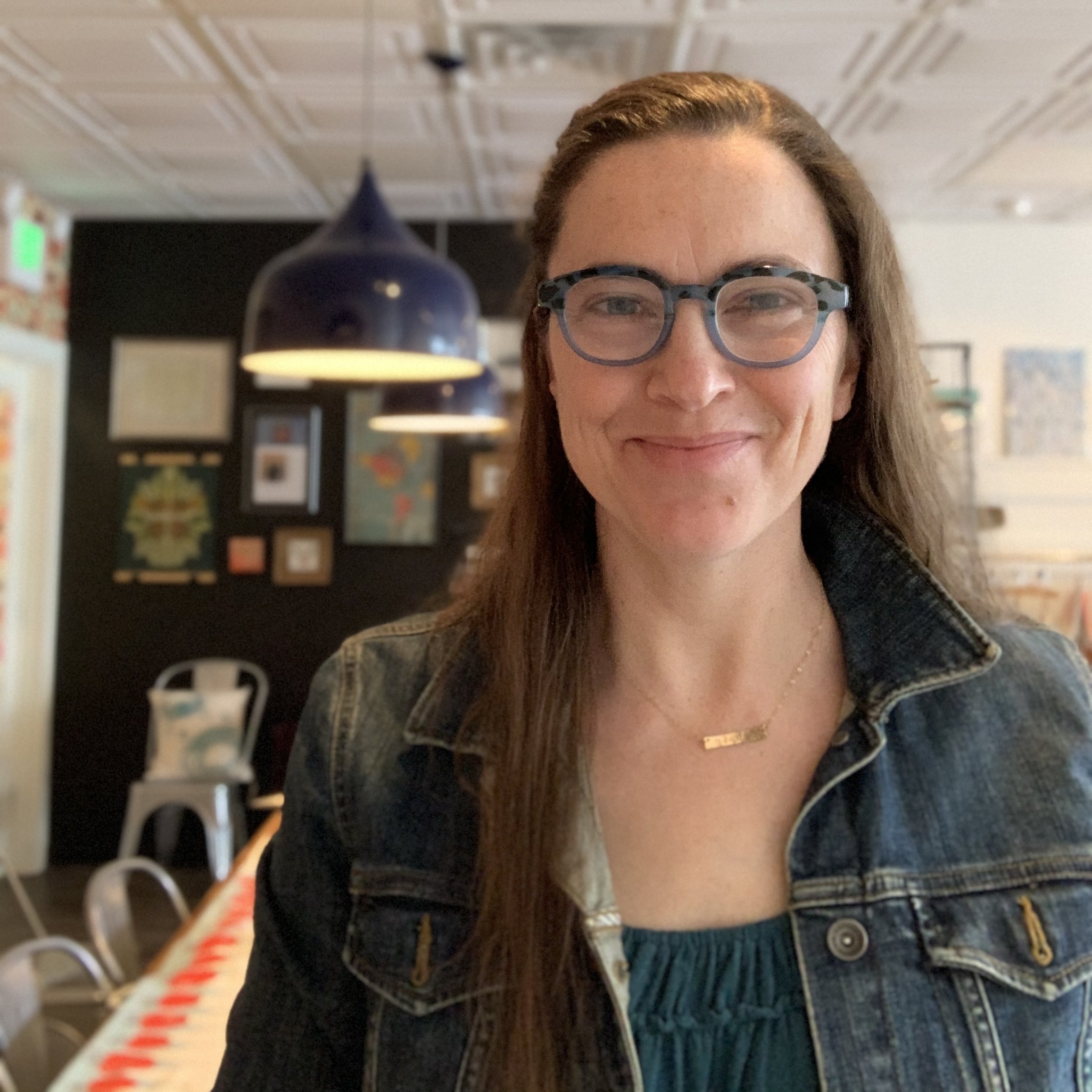 A woman with glasses and long brown hair smiling, wearing a denim jacket and a gold necklace, in a cozy indoor space decorated with framed art on the wall.