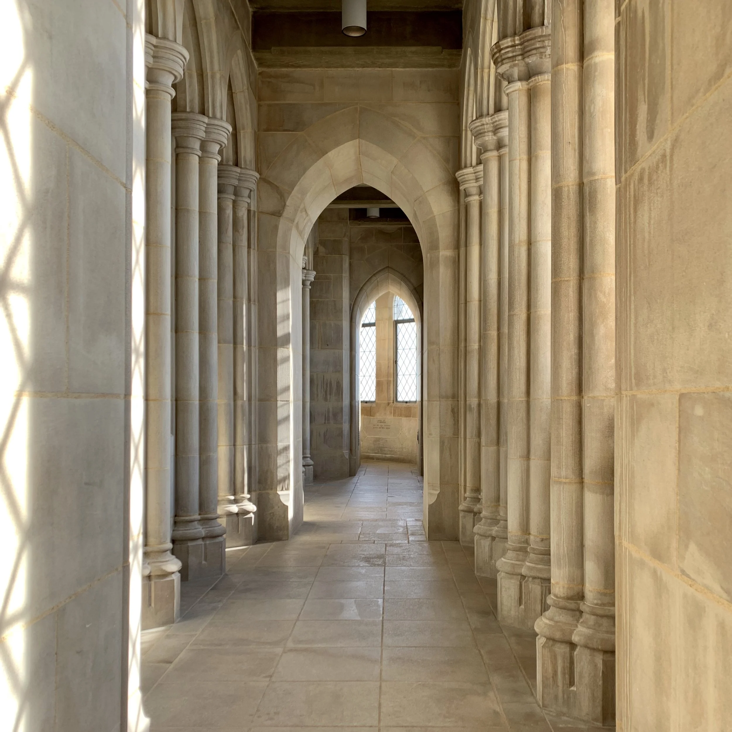 A corridor inside a stone building with arched windows and detailed columns, illuminated by natural sunlight.