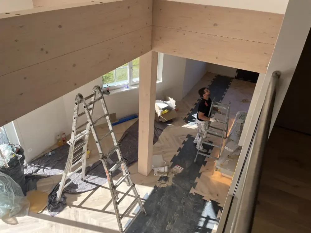 Innenaufnahme eines Hauses während der Renovierung. Es sind mehrere Leitern, Arbeitsmaterialien und eine Person zu sehen, die an einem Dach arbeitet, das mit Holzpaneelen verkleidet wird.