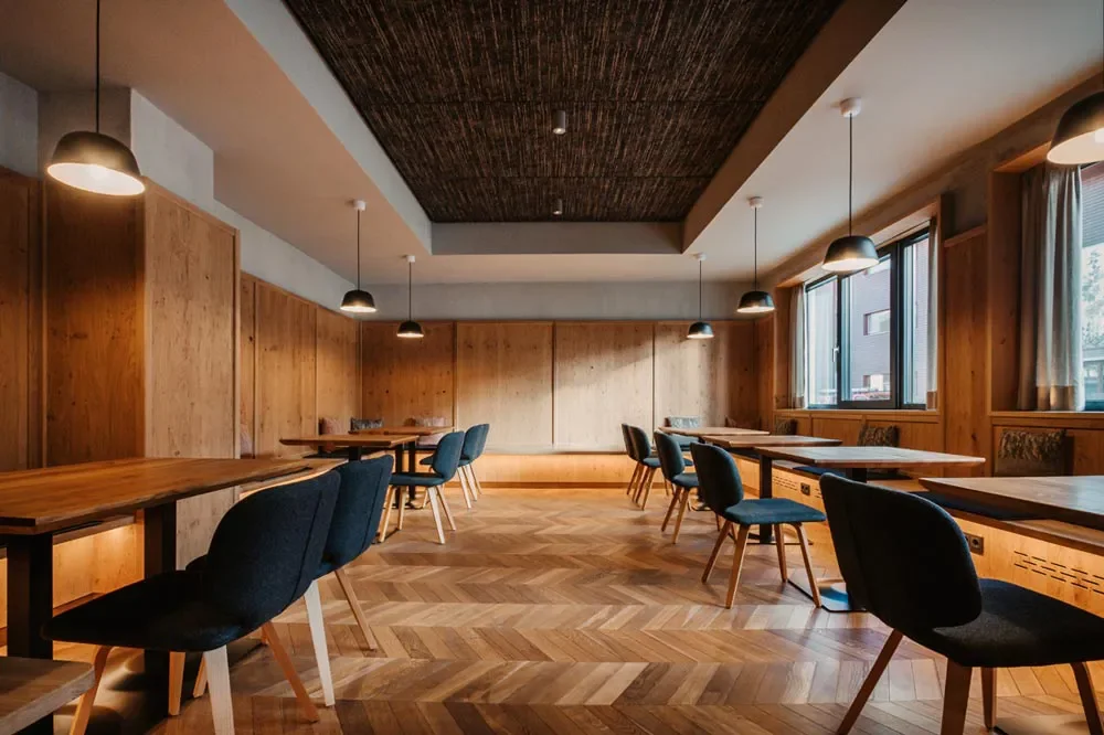 Ein modern möbliertes Esszimmer mit Holzpanelen, Holztisch und schwarzen sowie blauen Stühlen, große Fenster mit Vorhängen, Deckenleuchten, Holzparkettboden.