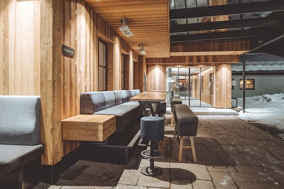 Innenaufnahme eines Holz-Restaurants oder Cafés, mit Sitzbänken, Tischen und Stühlen, bei Nacht. Im Hintergrund sind große Fensterscheiben und Schnee draußen sichtbar.