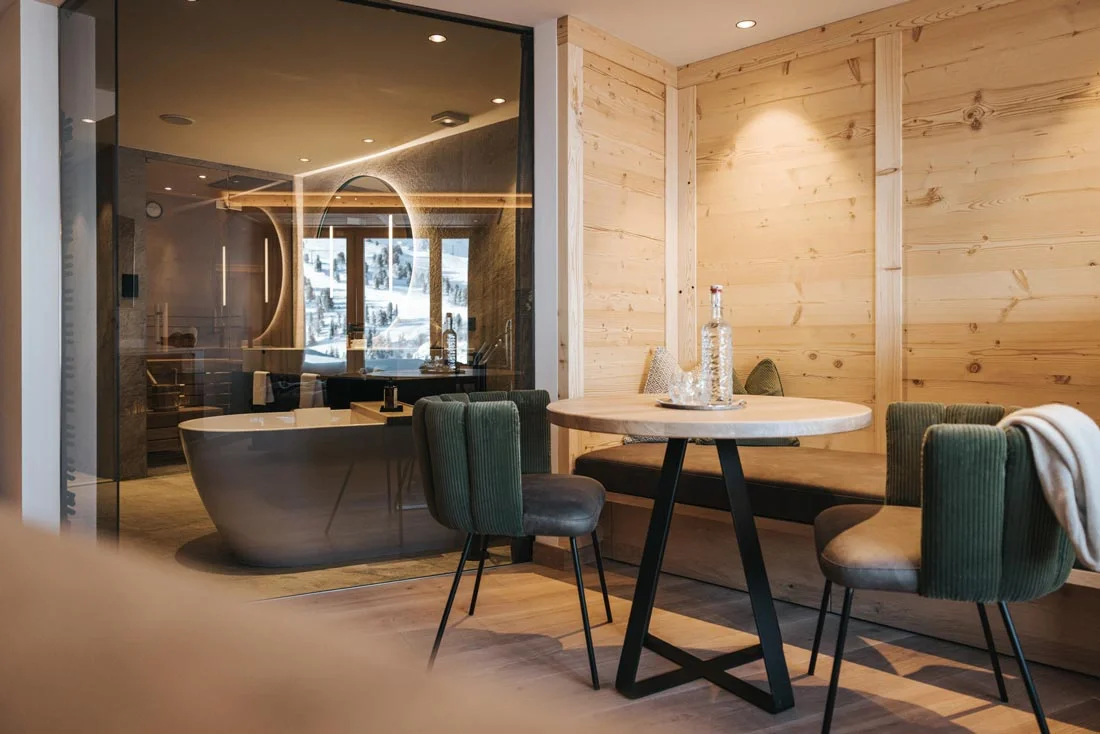 Ein modernes, gemütliches Wohnzimmer mit Holzvertäfelung, einem runden Esstisch mit Stühlen und einer Badewanne im Hintergrund, durch eine Glastür getrennt.