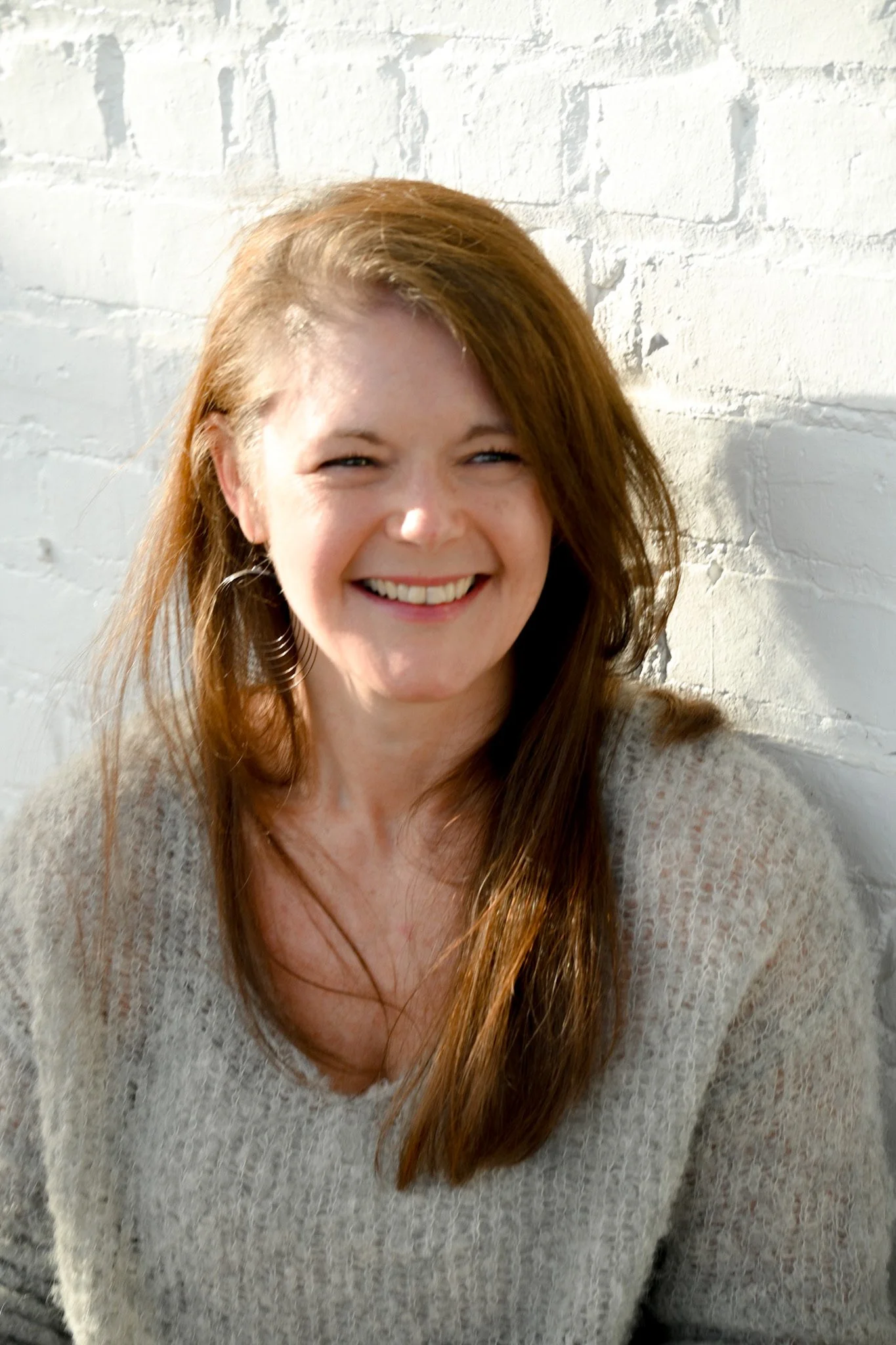 Close-up of a smiling woman with long, red hair wearing a beige sweater against a white brick wall.
