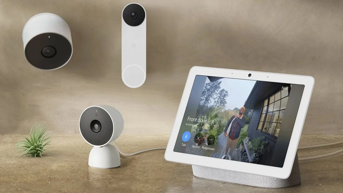 Smart home security system with a Google Nest Hub displaying a front door surveillance feed. Includes a white Nest Cam, a Nest Doorbell, and a second small security camera on a countertop with a small green plant.
