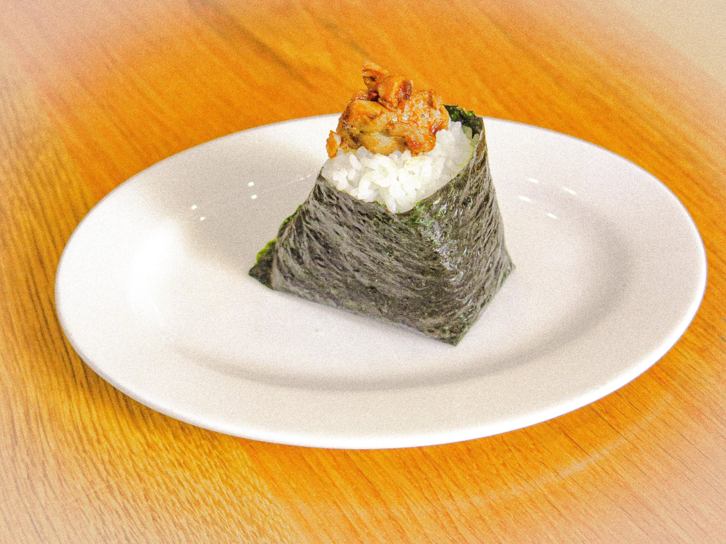 A piece of sushi with rice and seaweed wrapping, topped with cooked eel or fish on a white plate placed on a wooden surface.