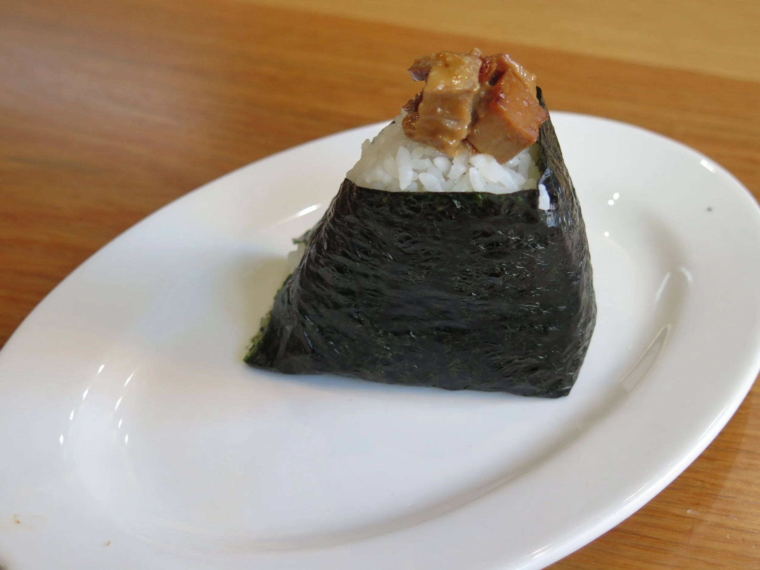 A rice ball wrapped in seaweed with a topping of cooked meat pieces on a white plate.