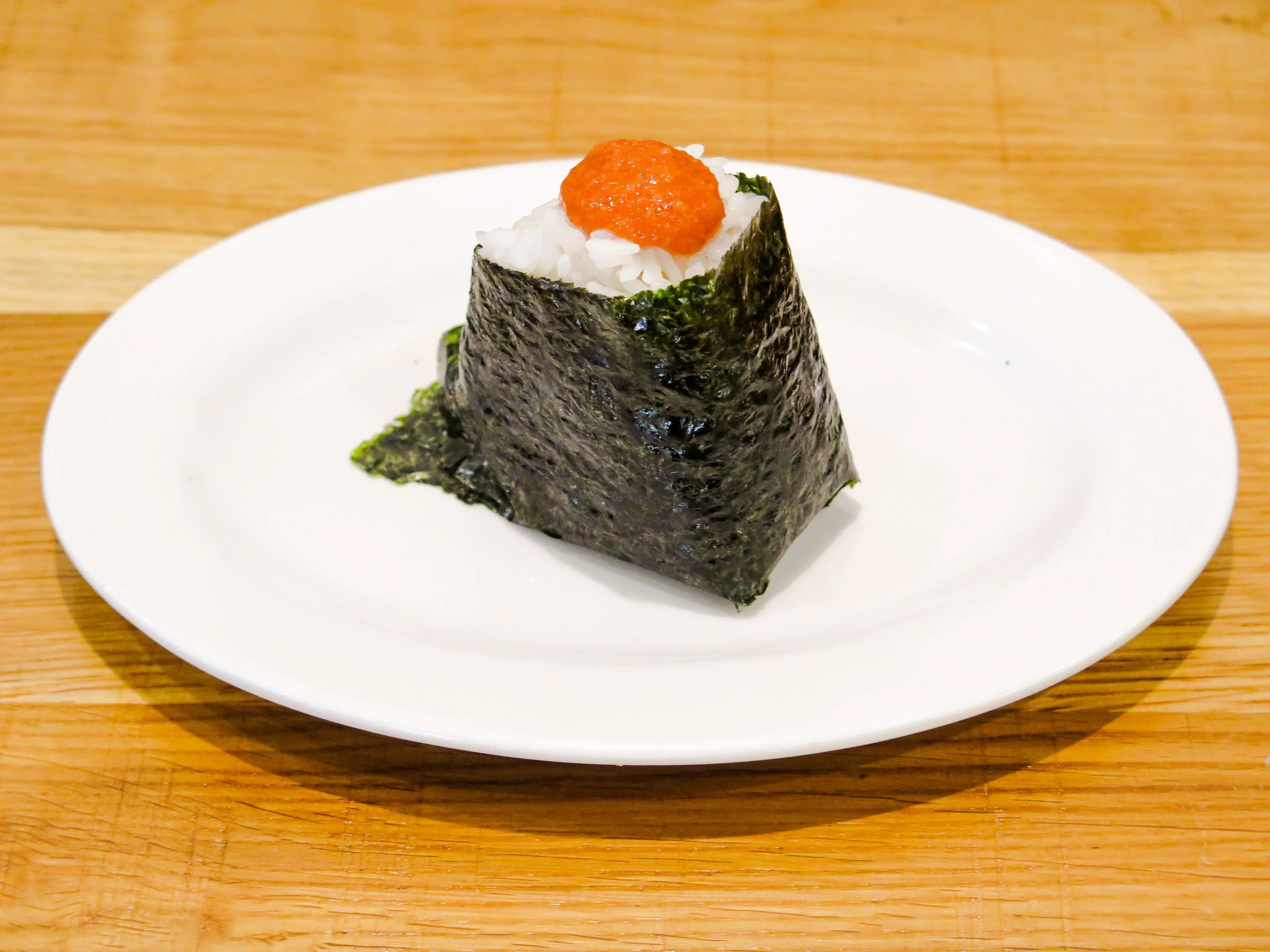 A single piece of sushi on a white plate, wrapped in nori seaweed, with white rice and orange sauce on top, placed on a wooden surface.