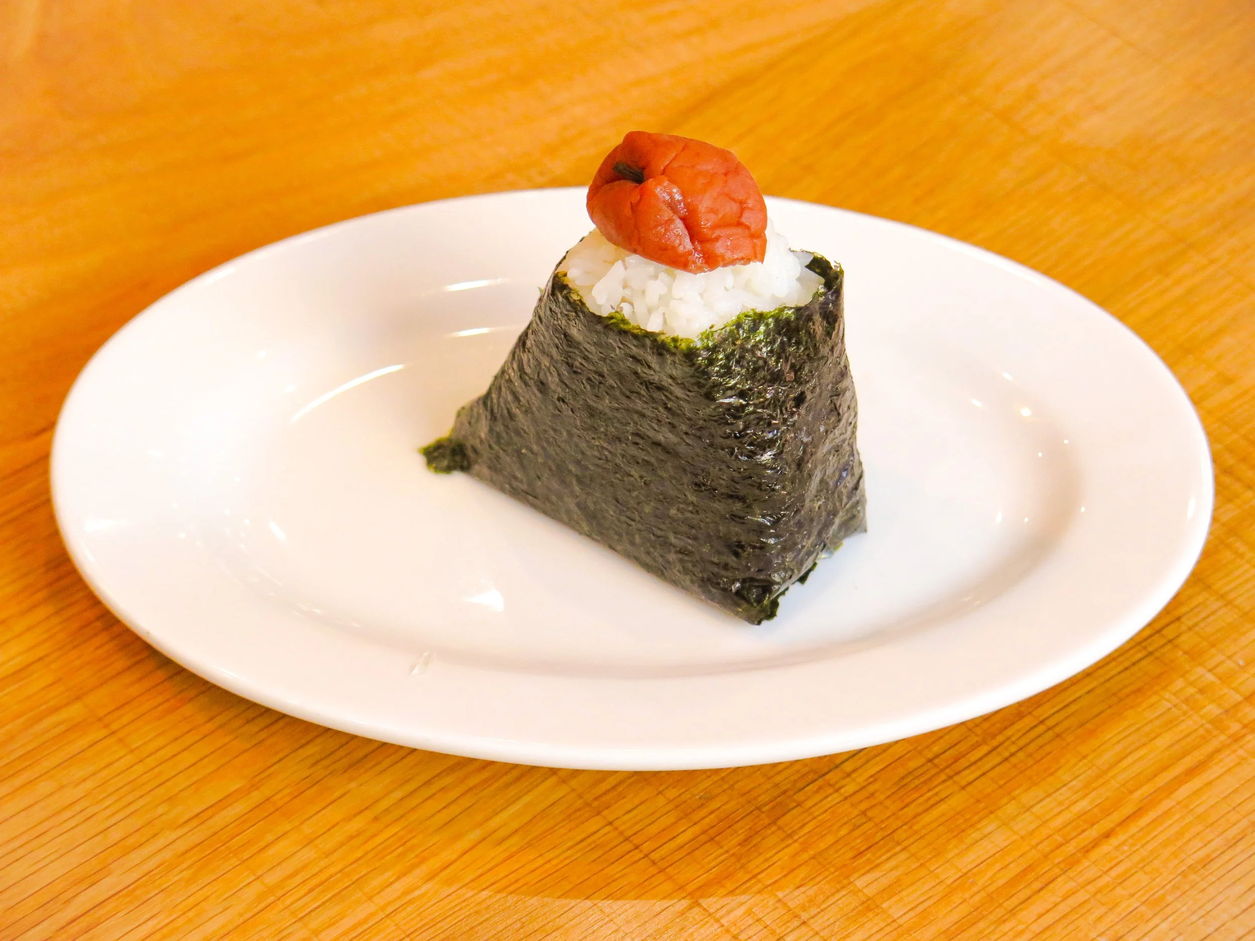 A piece of onigiri rice ball wrapped in nori seaweed, topped with a pickled plum and a small pickled vegetable.