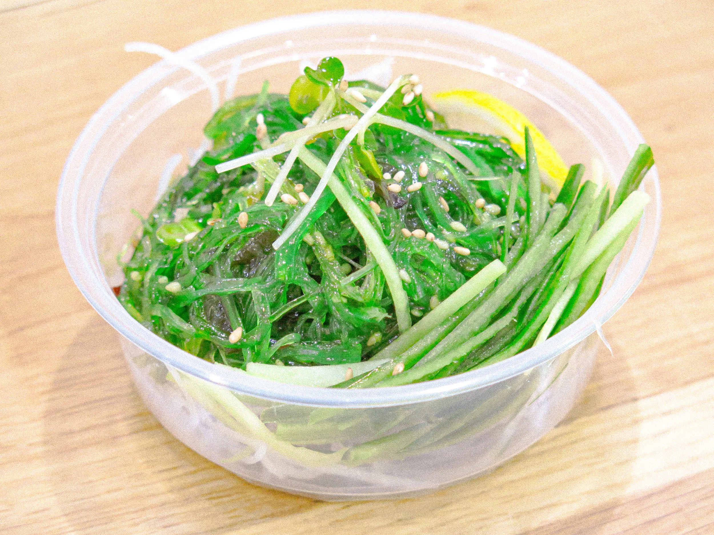 Seaweed salad garnished with sesame seeds and sliced cucumber in clear plastic container on wooden surface.