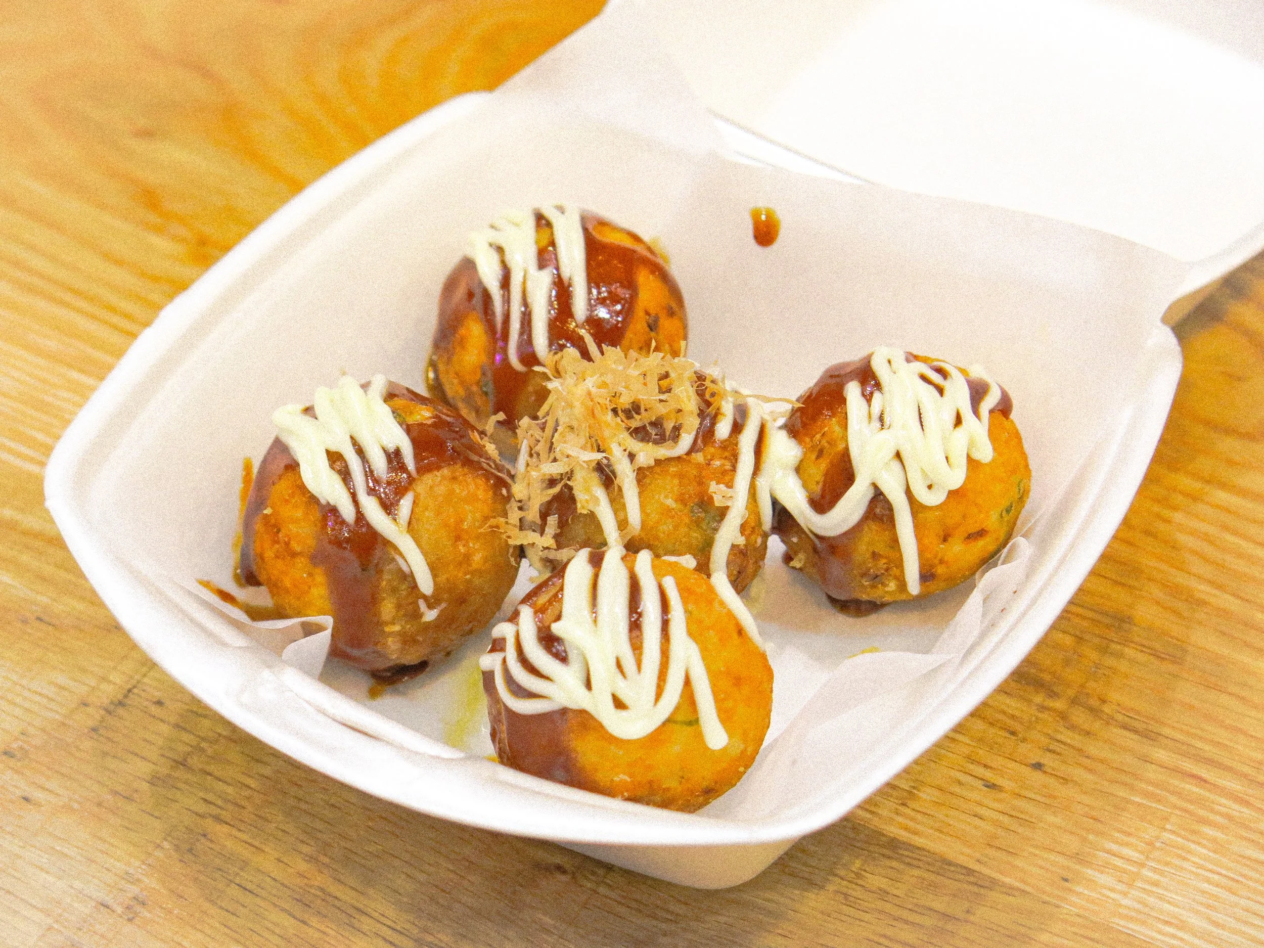 A white takeout container with five takoyaki balls topped with mayonnaise, sauce, and bonito flakes on a wooden surface.