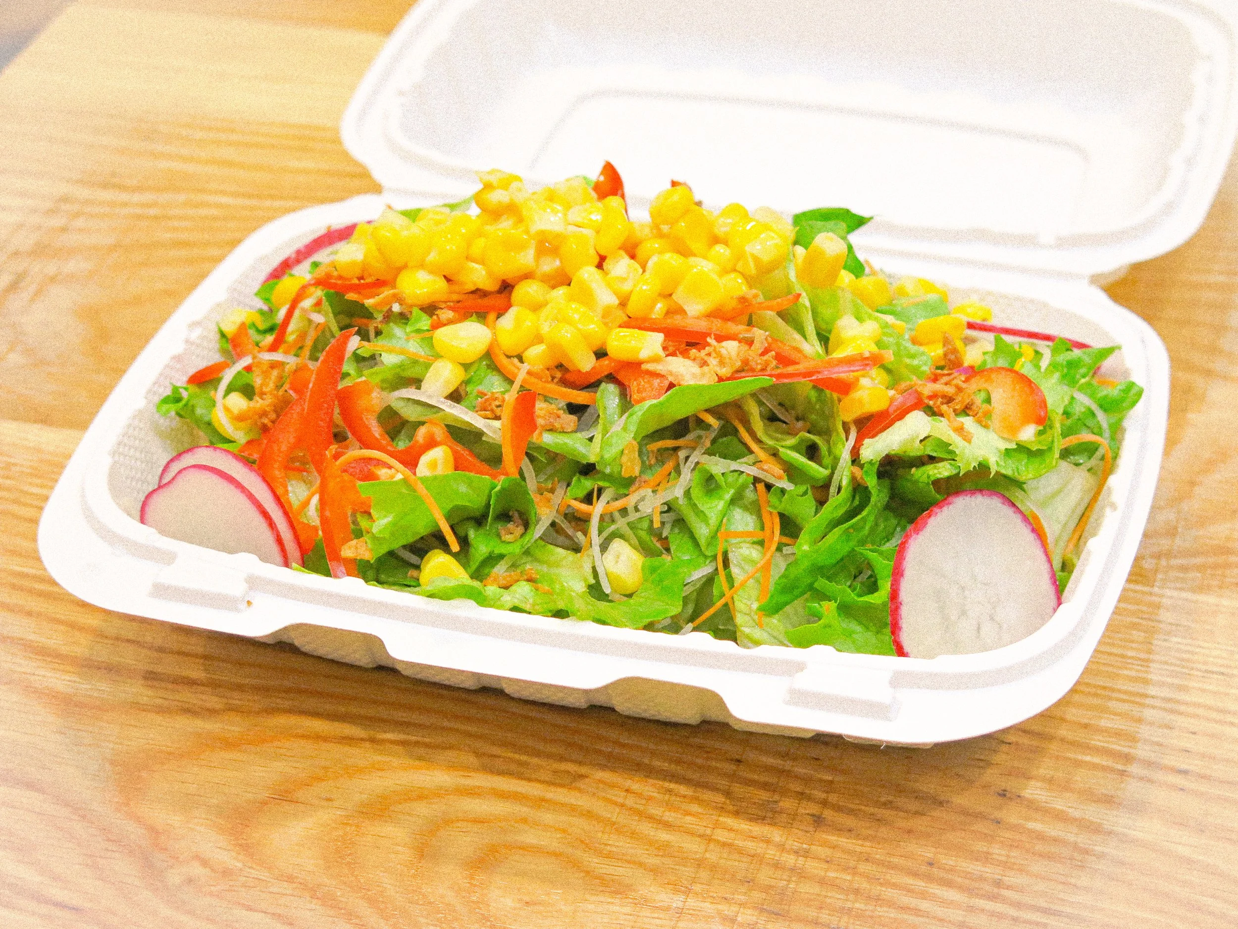 Salad with lettuce, radishes, shredded carrots, corn, and green onions in a white takeout container on a wooden surface.