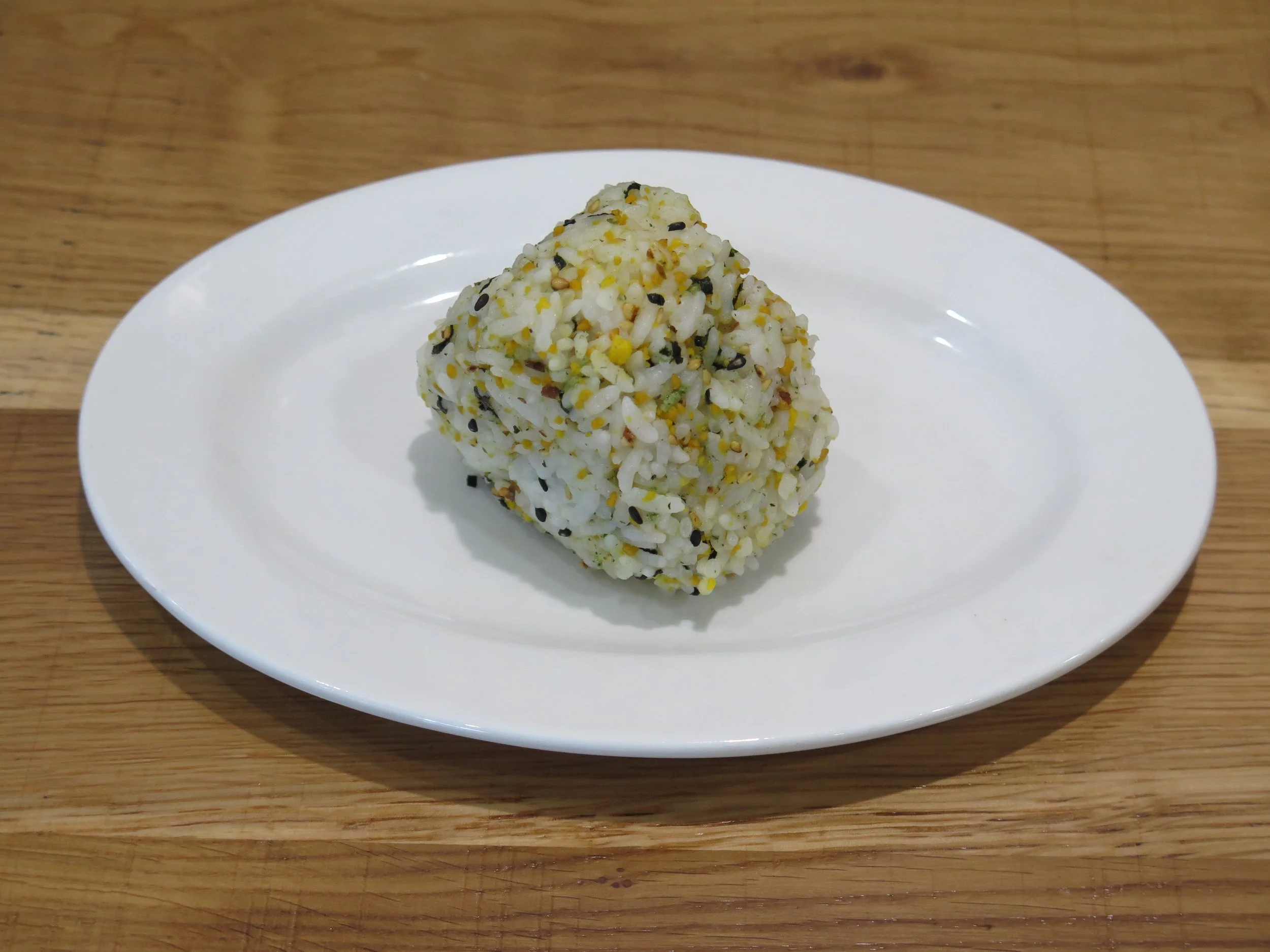 A rice ball coated with sesame seeds and spices on a white oval plate, placed on a wooden surface.