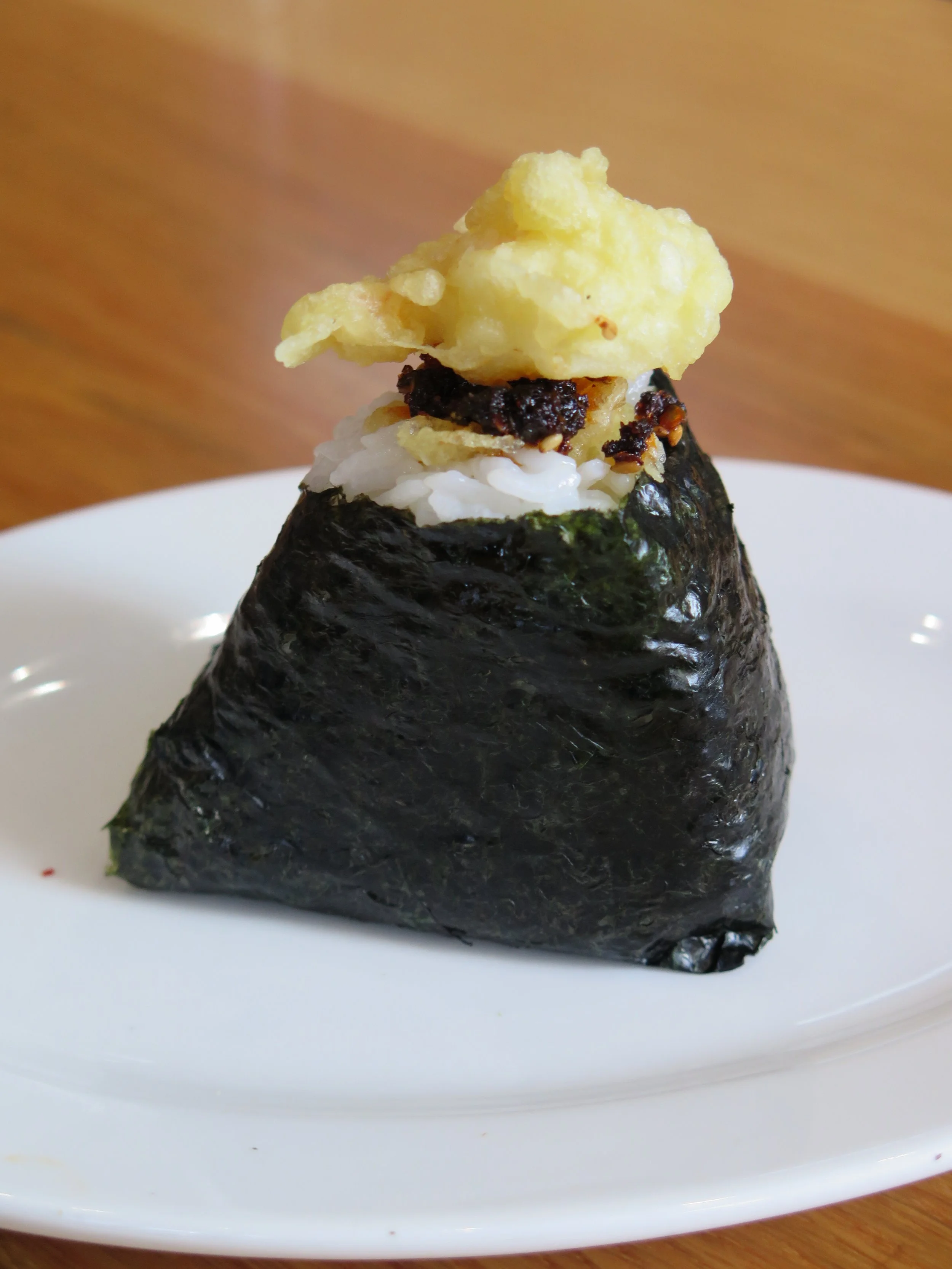 A Japanese onigiri rice ball wrapped in seaweed with tempura topping on a white plate.