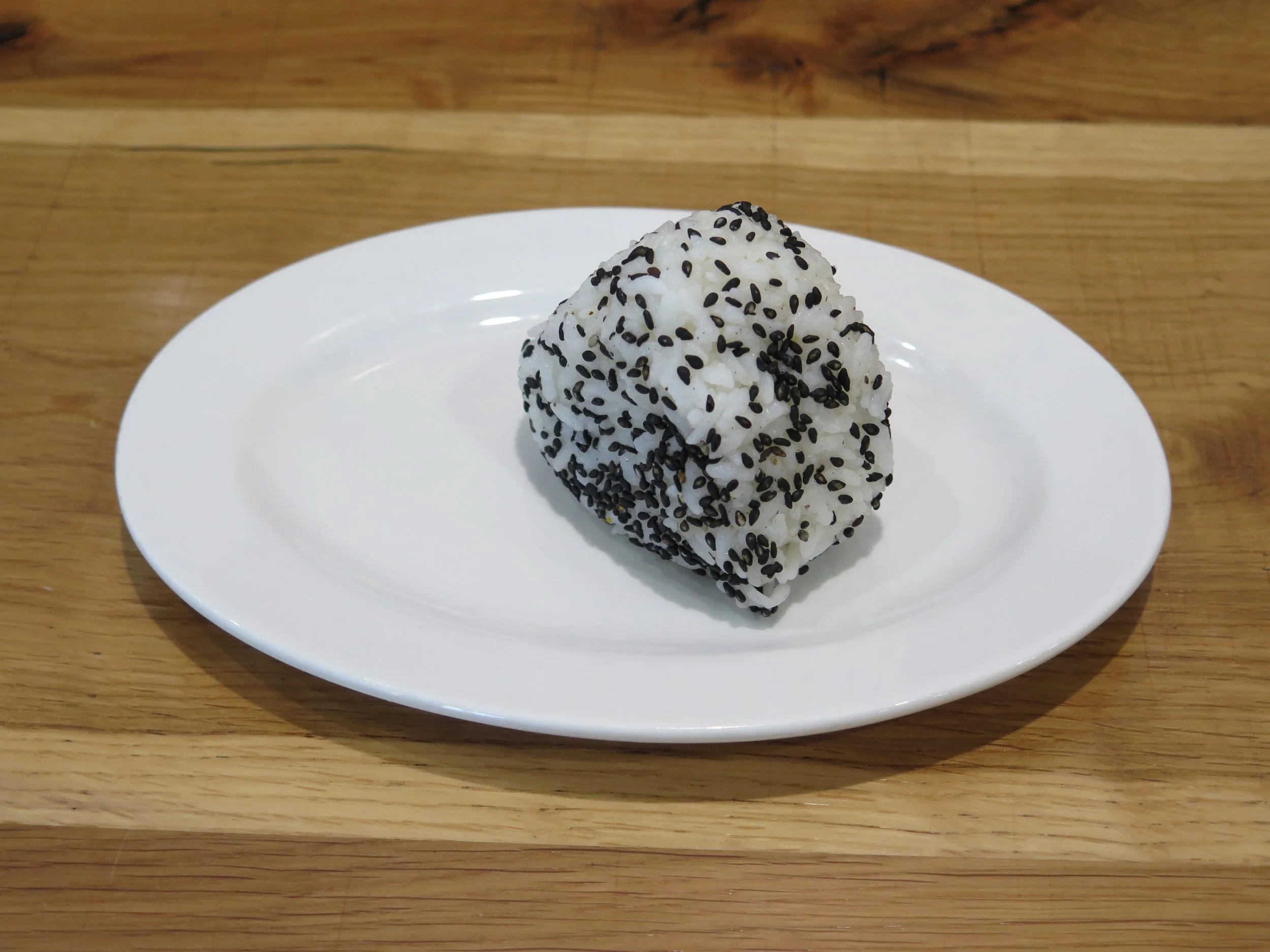 A rice ball coated in black sesame seeds on a white oval plate, placed on a wooden surface.
