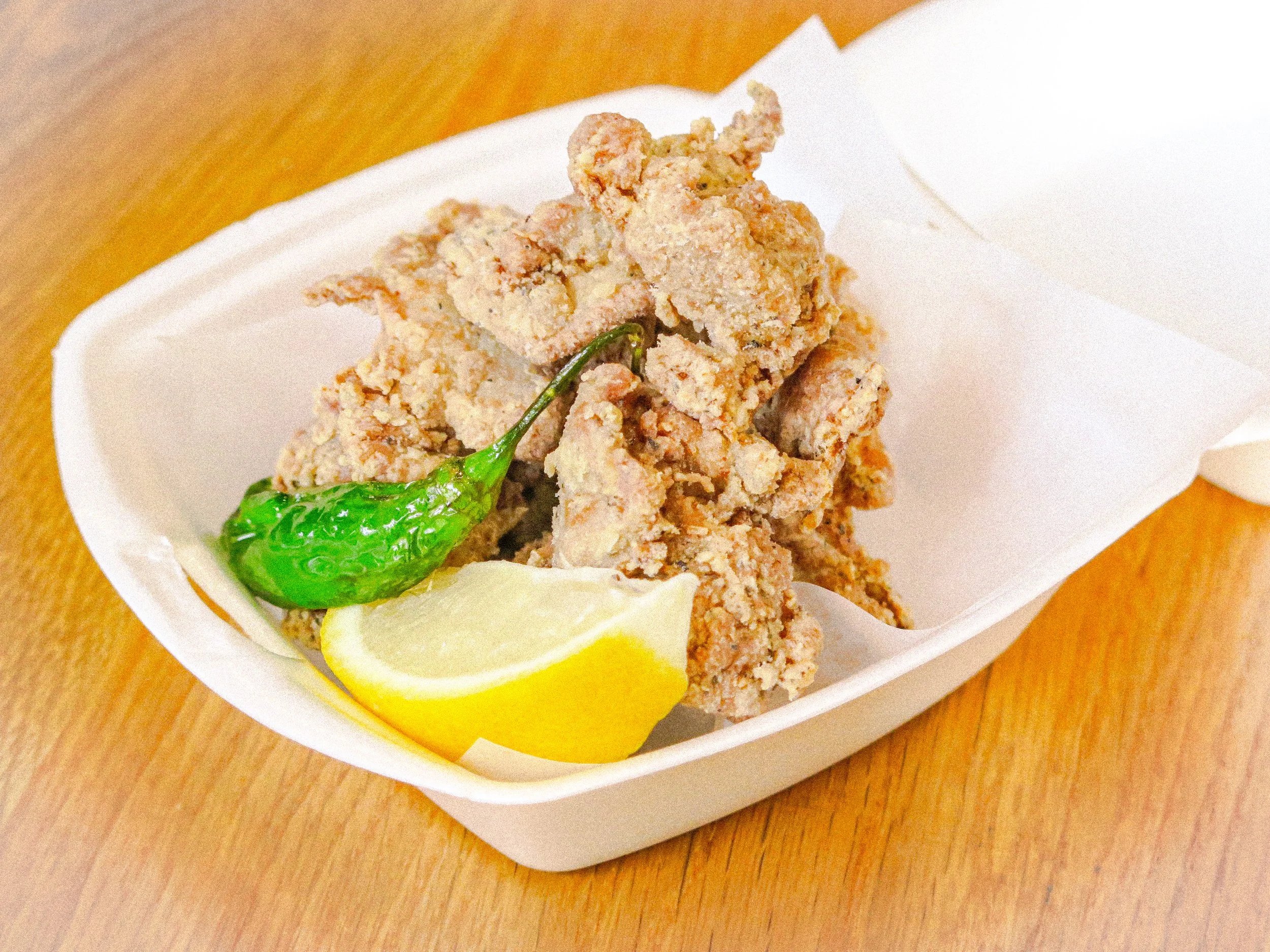 Fried chicken pieces garnished with a green pepper and lemon wedge in a white paper takeout box on a wooden surface.