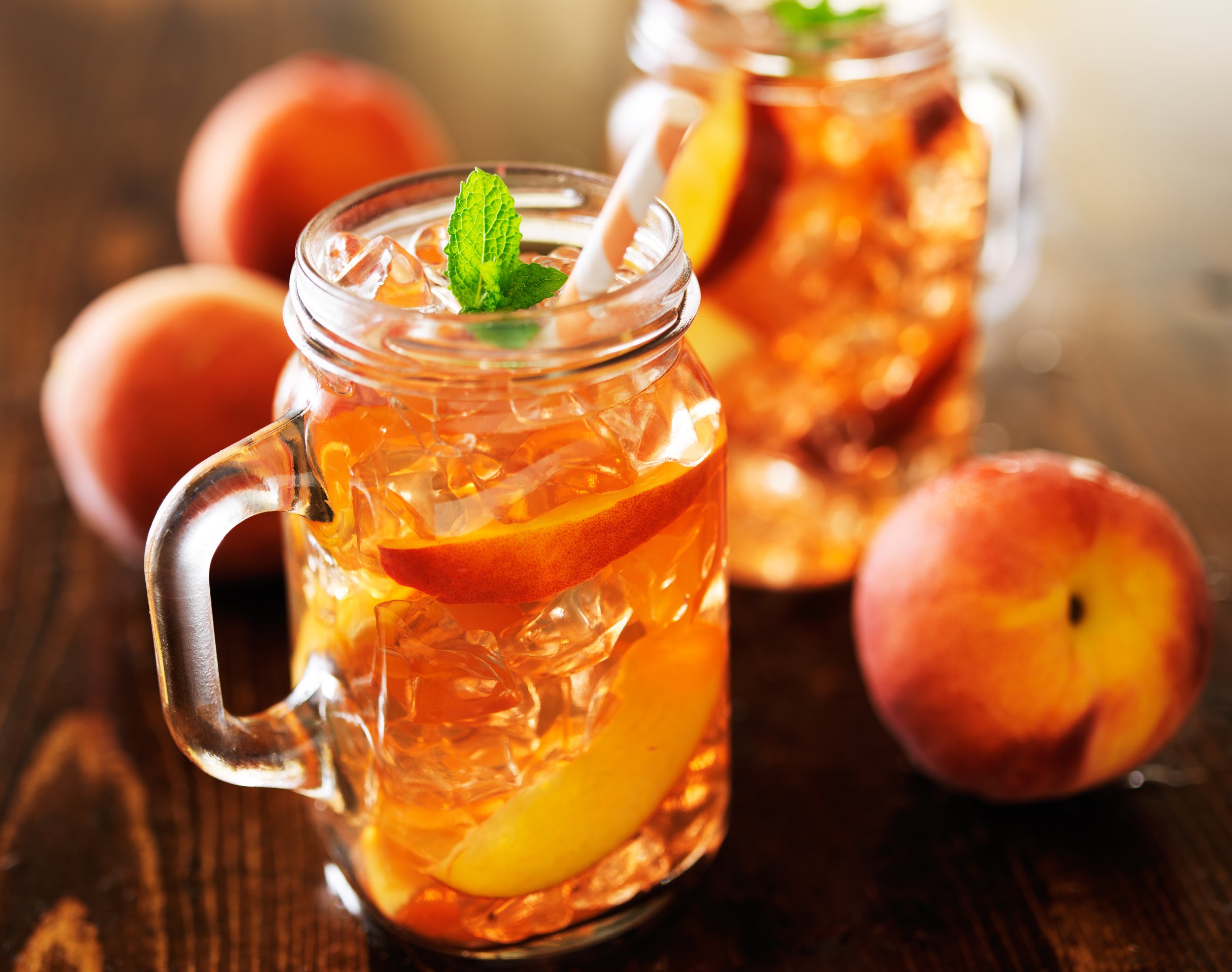 Glass jars filled with peach iced tea, garnished with mint and lemon slices, surrounded by fresh peaches on a wooden surface.