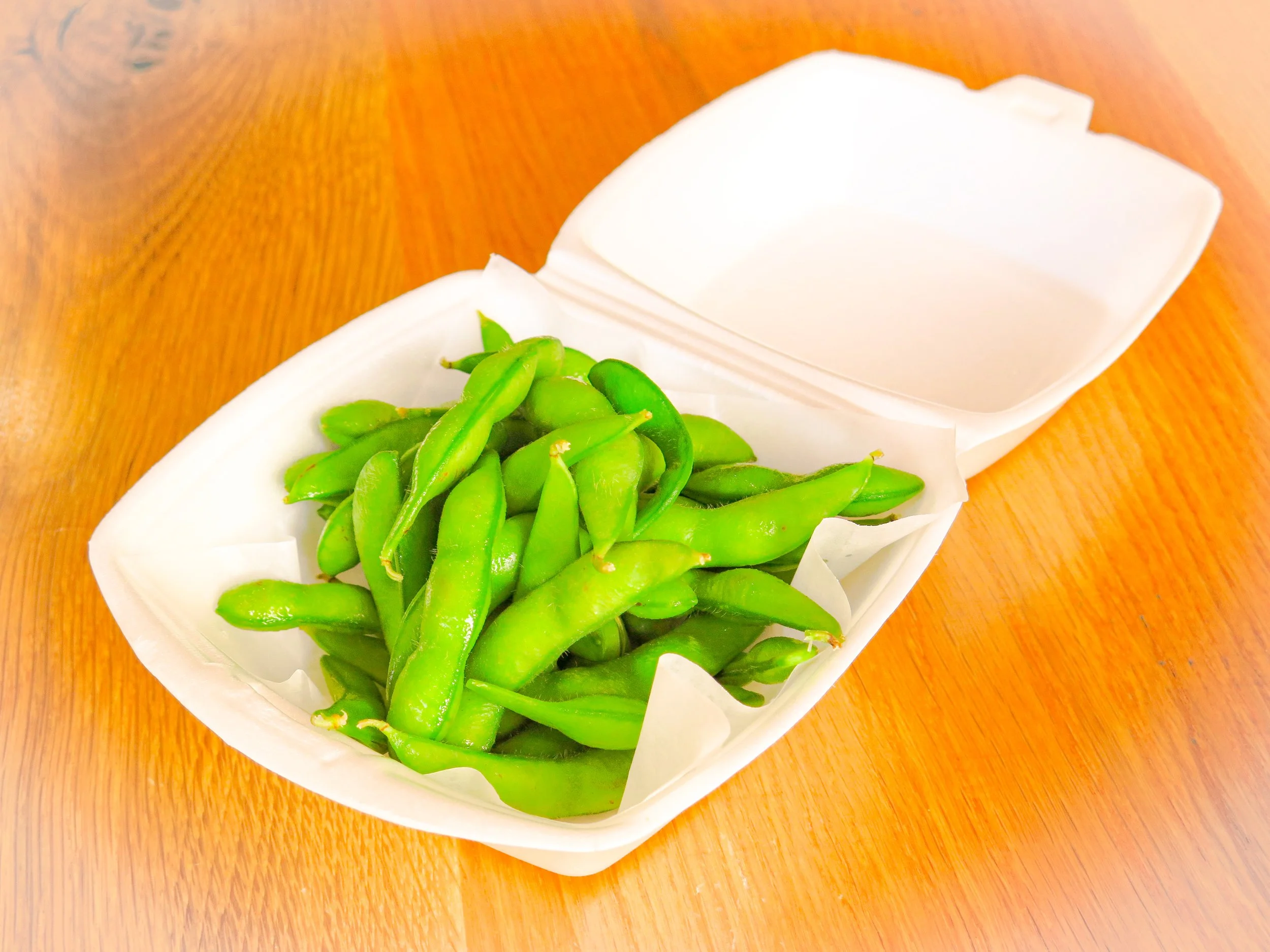 Green edamame beans in a white foam takeout container on a wooden table.