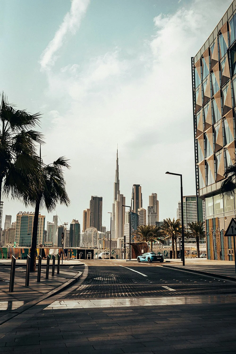 Skyline view of Dubai with the Burj Khalifa in the center, modern buildings, palm trees in the foreground, and a car parked on the street.