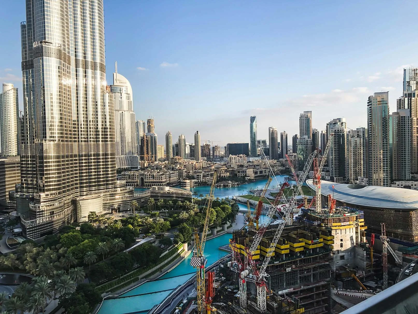 A cityscape with numerous modern skyscrapers, construction cranes, and a river flowing through the urban area, under a blue sky.