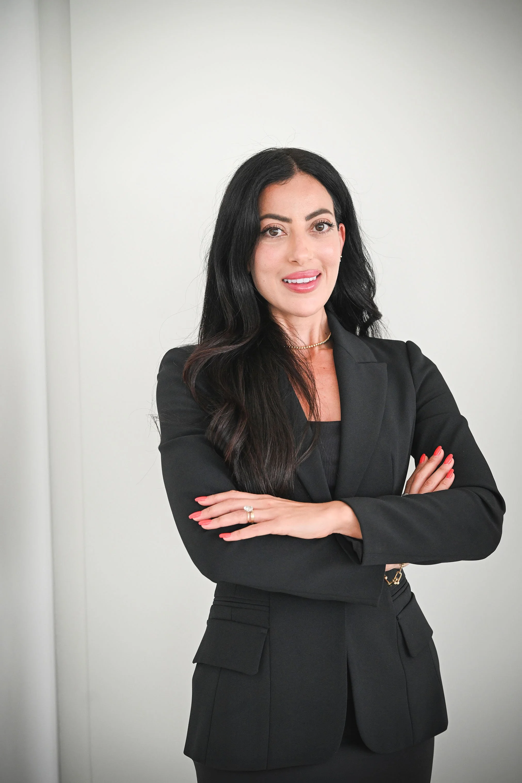 A woman with long black hair, dressed in a black suit, standing with arms crossed against a plain white wall.