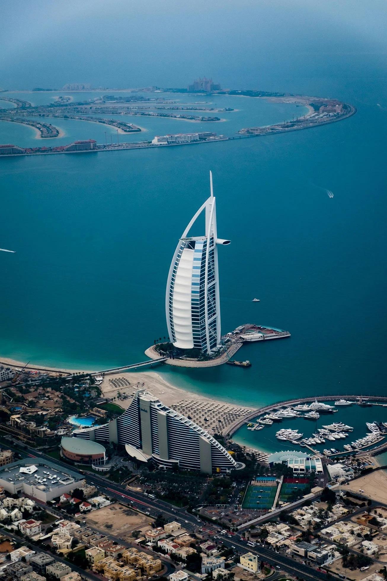 Aerial view of the Burj Al Arab hotel, shaped like a sail, located on a man-made island off the coast of Dubai, United Arab Emirates, with surrounding water, marina, and cityscape.