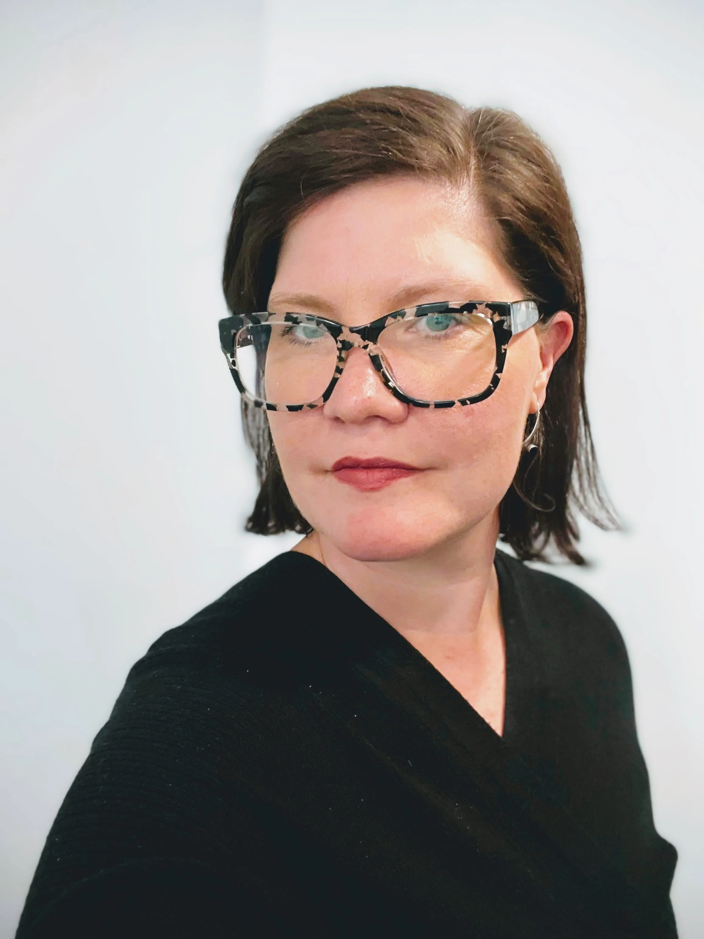 A woman with short brown hair, wearing glasses with a black and white patterned frame, is captured in a close-up photo against a off-white background. She is wearing a black top.