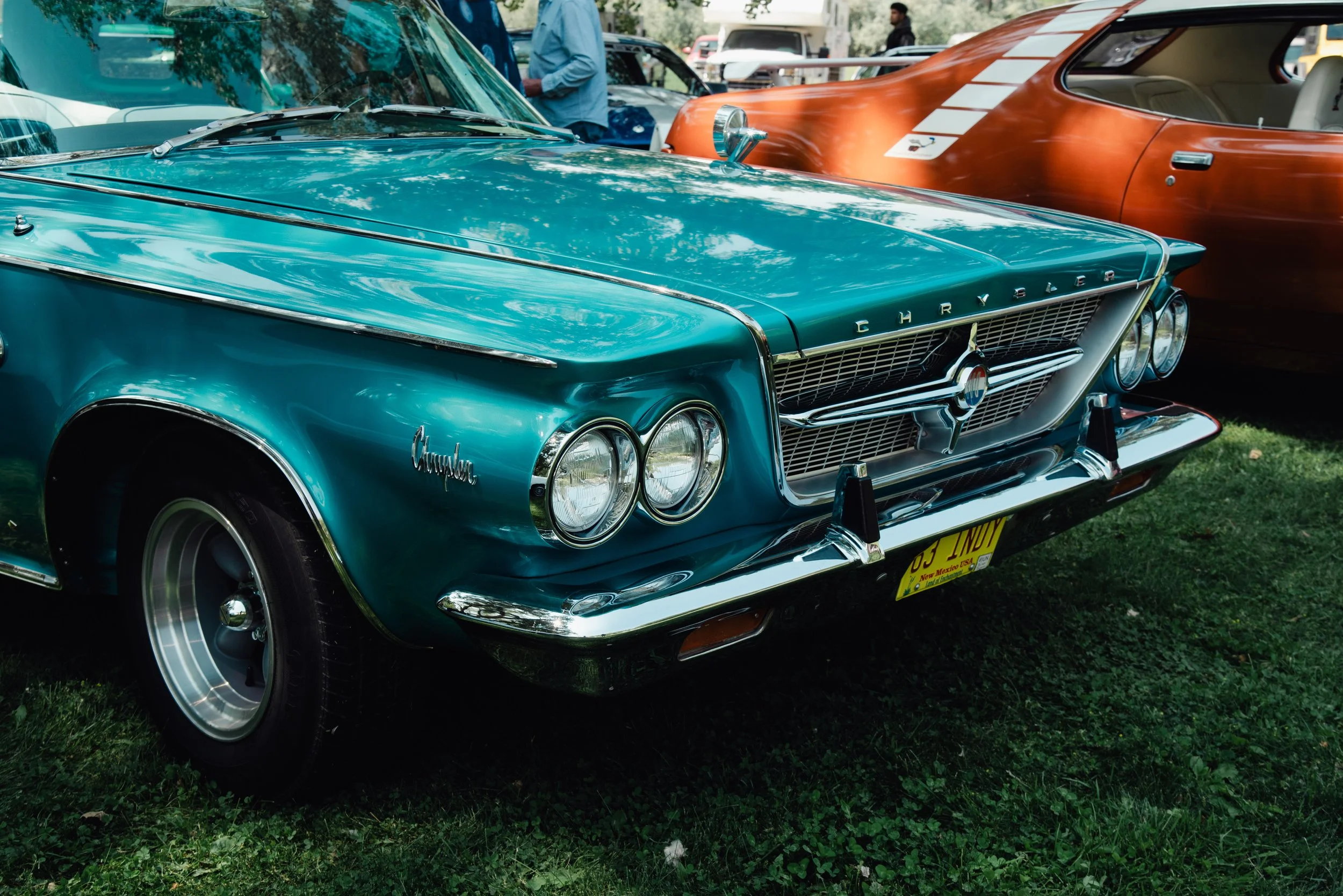A vintage teal Chevrolet Corvair car parked outdoors at a car show, with other classic cars and people visible in the background.