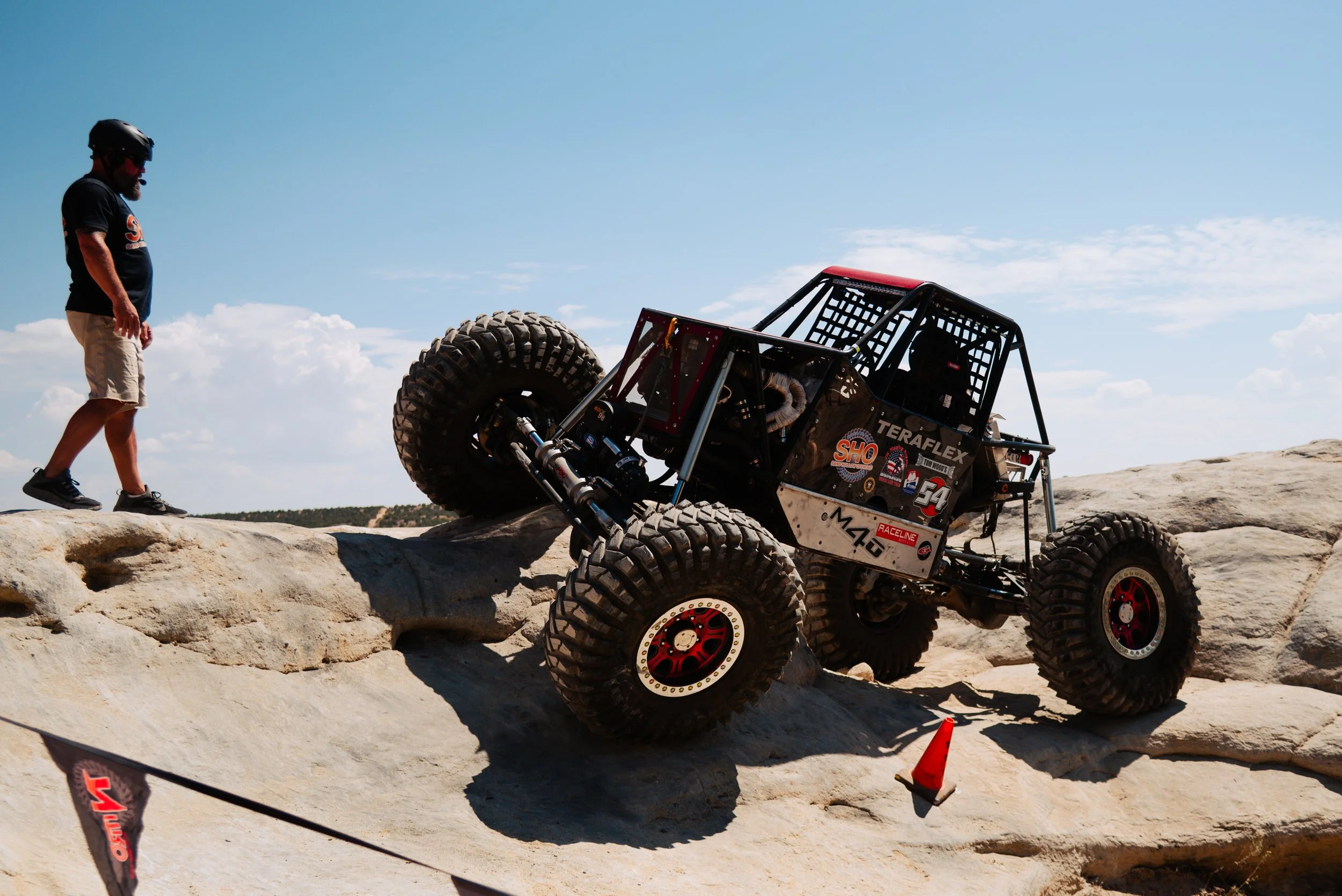 An off-road rock crawler in a black roll cage and large tires, climbing over rocks during a competition, with a man in cycling gear observing nearby, under a blue sky.