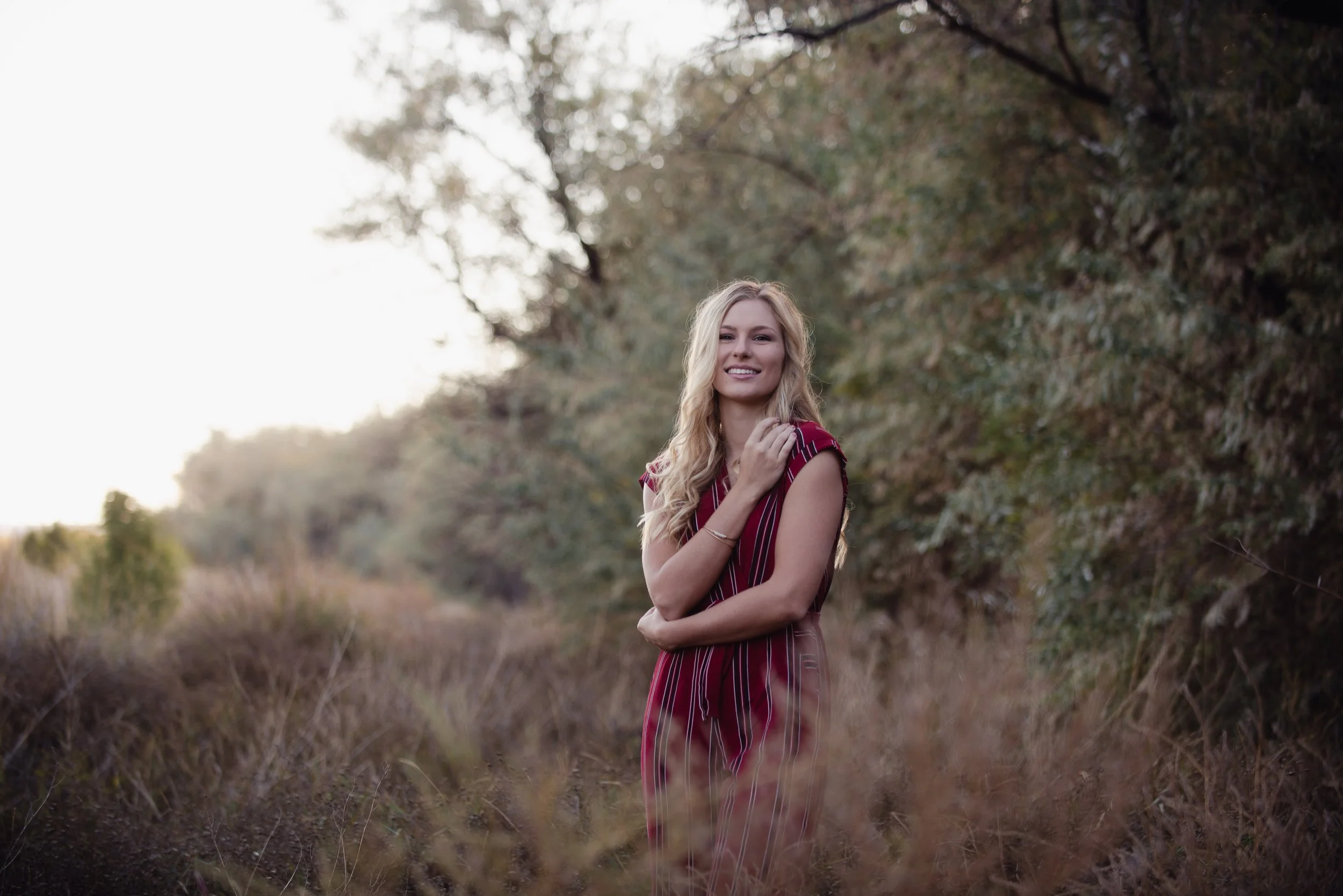 A woman with blonde hair smiling, standing in a field with tall grass and trees in the background during sunset.