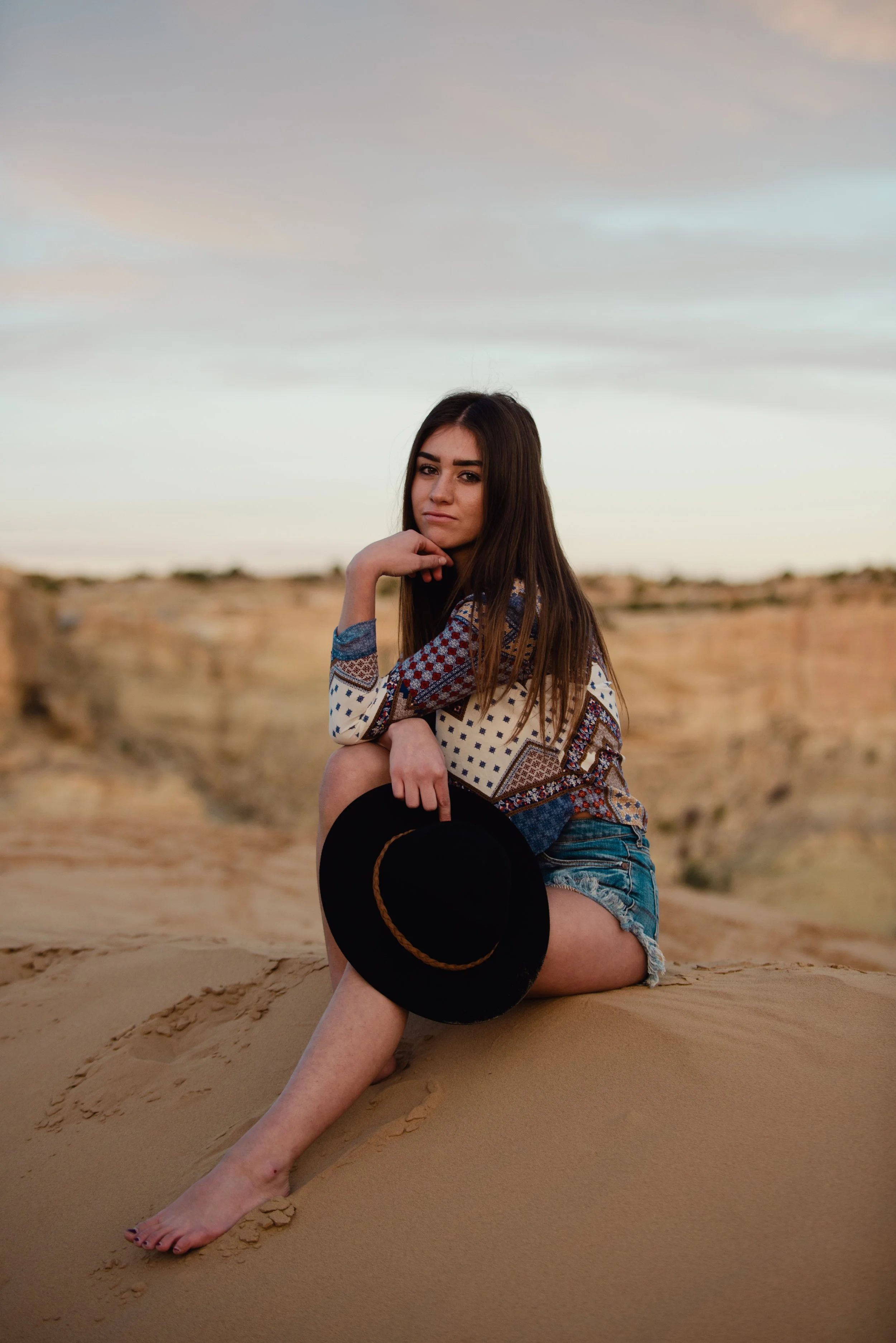 A young woman with long brown hair sitting on sand dunes in a desert landscape at sunset, holding a black hat with a brown band, wearing a patterned long-sleeve shirt and denim shorts.