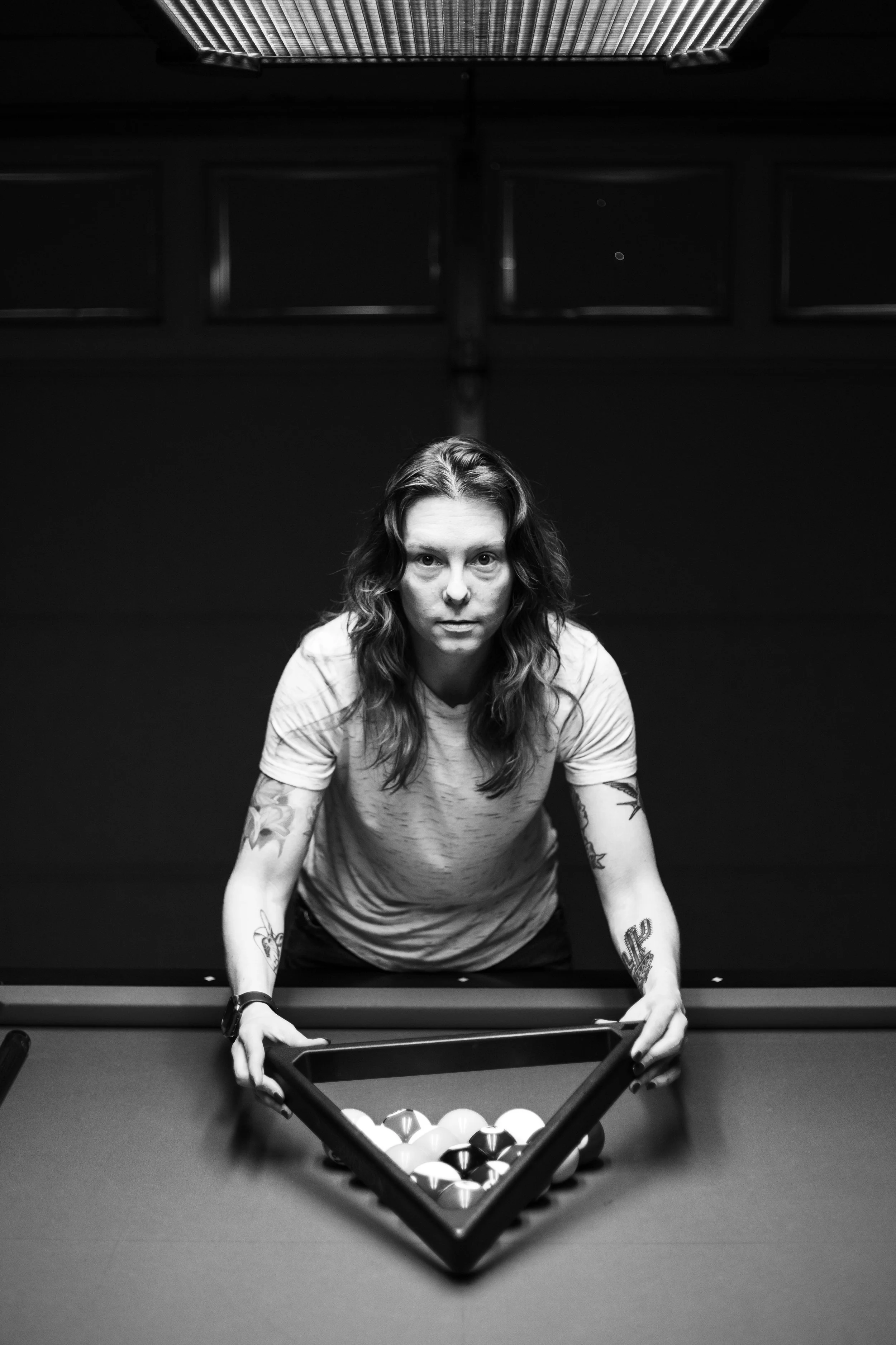 A woman with wavy hair and tattoos on her arms leaning over a pool table, holding the racking triangle, with pool balls arranged in a triangle on the table, viewed from above in black and white.