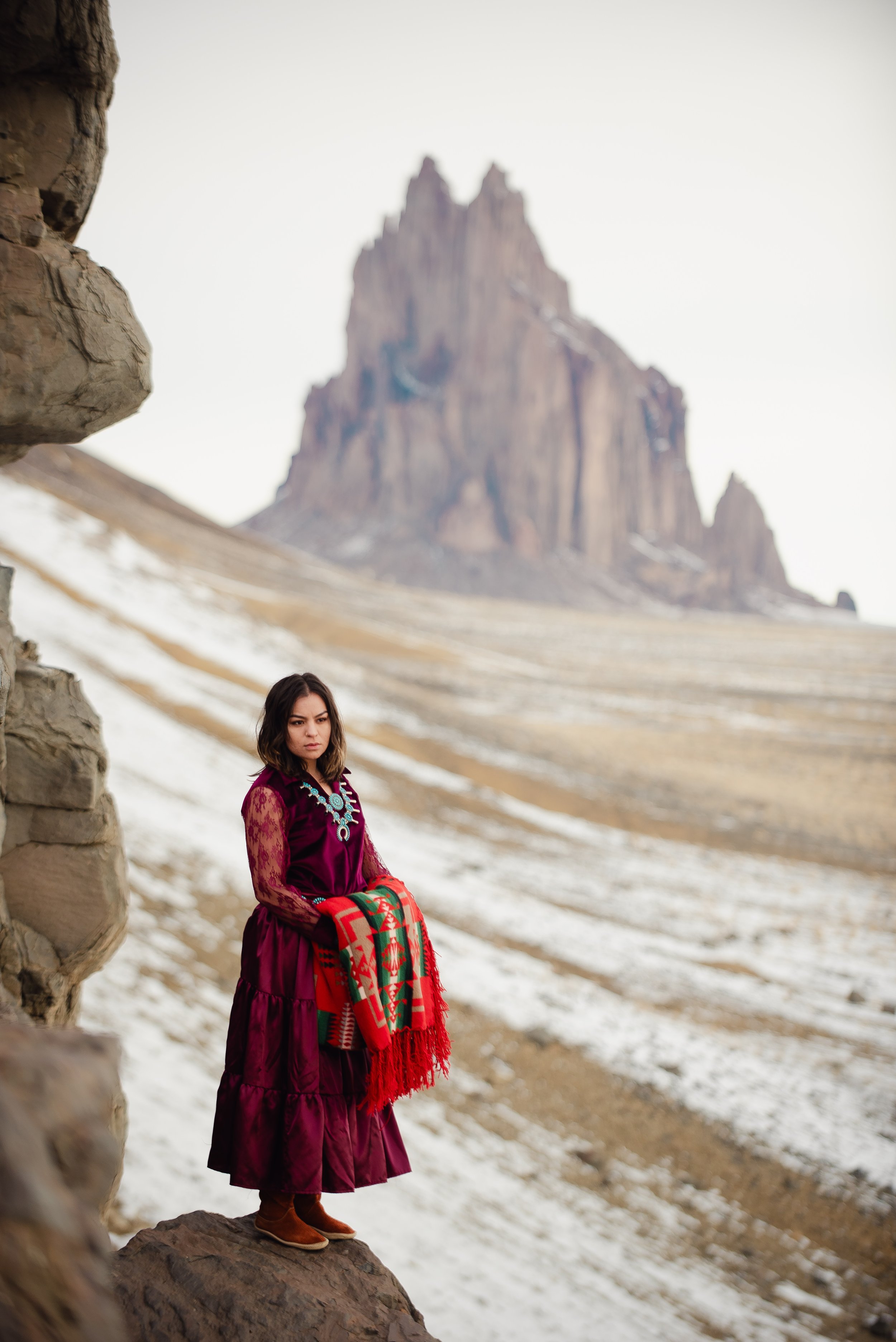 A woman in a purple dress and brown boots holding a colorful blanket stands on a rock in a mountainous, snowy landscape with large rock formations in the background.