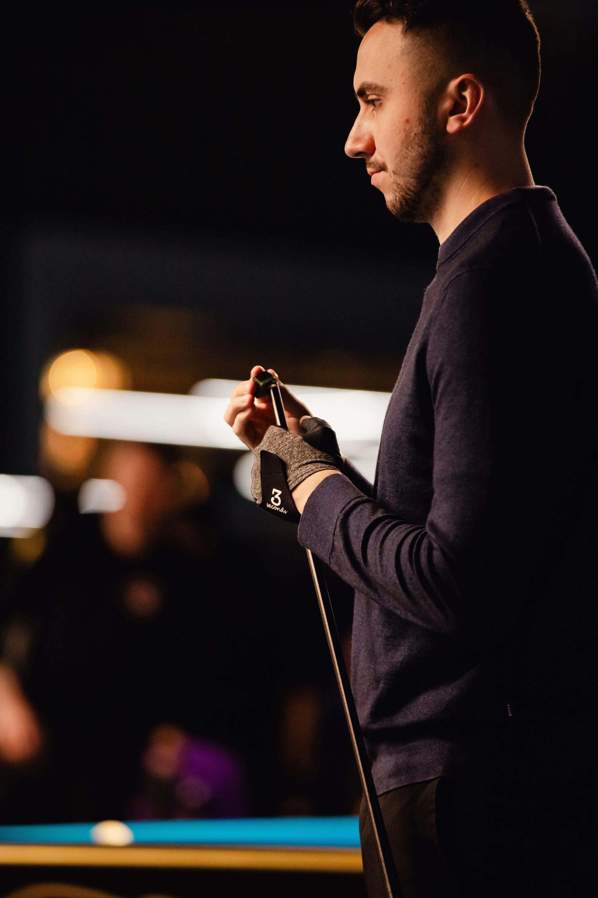 A young man in a dark long-sleeve shirt standing by a pool table, holding a pool cue, with a blurred background of people and lights.